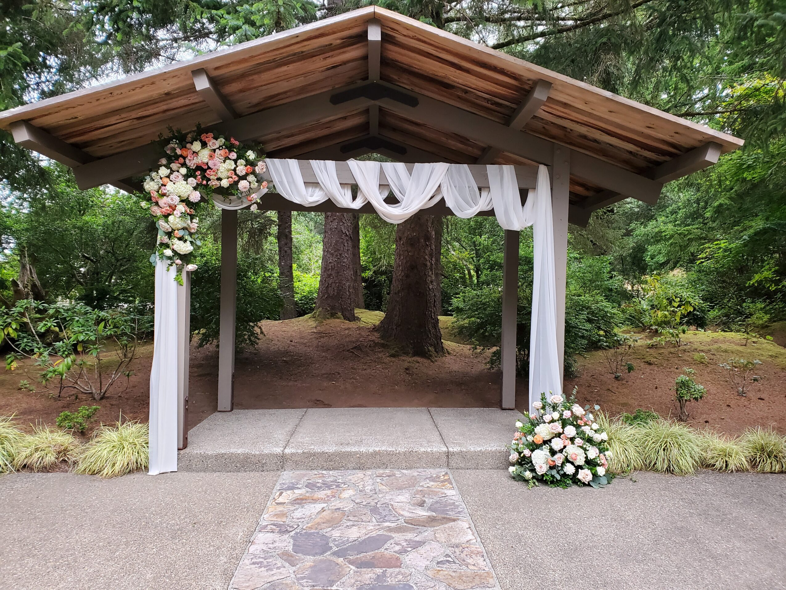 Wooden outdoor structure decorated with white fabric and floral arrangements, set in a green forested area.