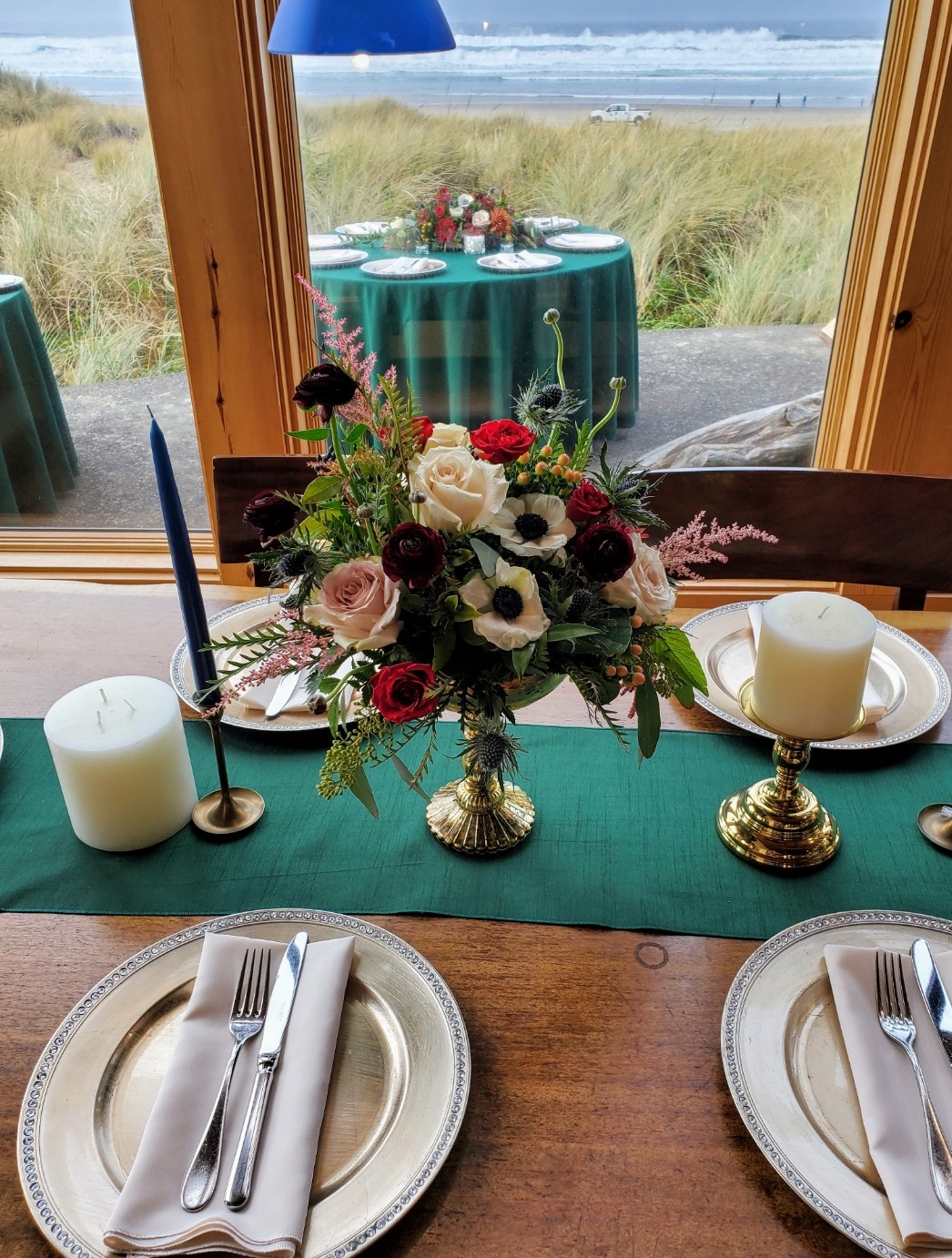 Table set with plates, silverware, candles, and a floral centerpiece, overlooking a beach scene through a window.