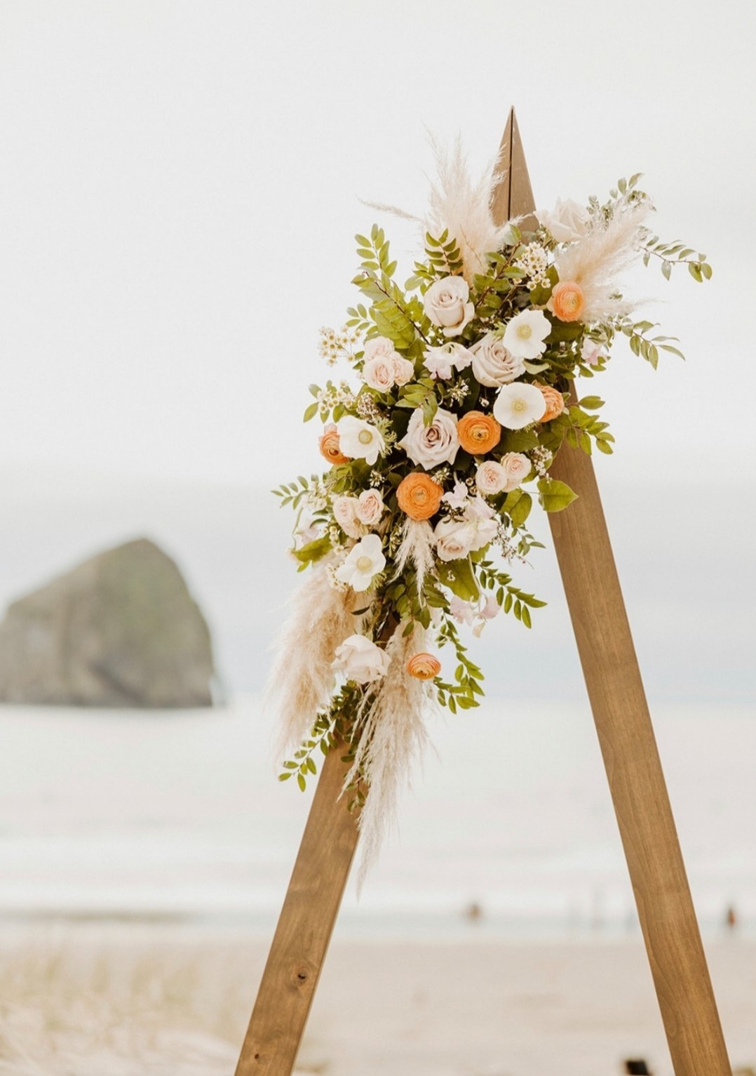 A floral arrangement on a wooden stand with white, peach, and green flowers, set against a beach background with a rock.