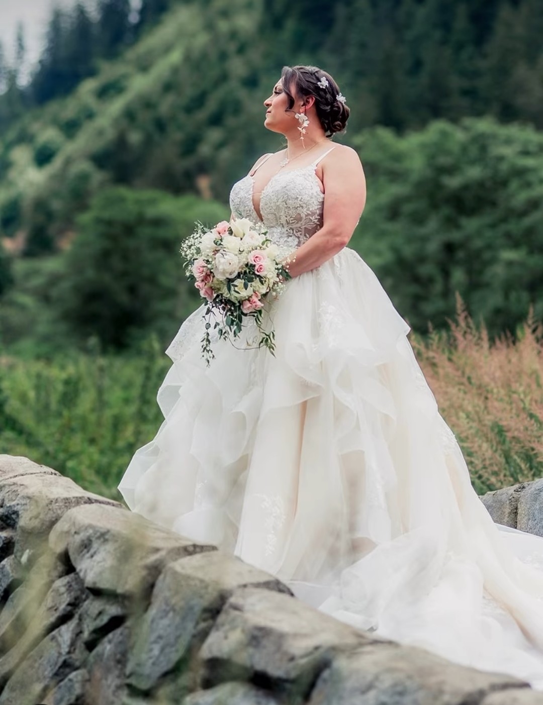 Bride in a white wedding gown holding a bouquet outdoors with trees and greenery in the background.