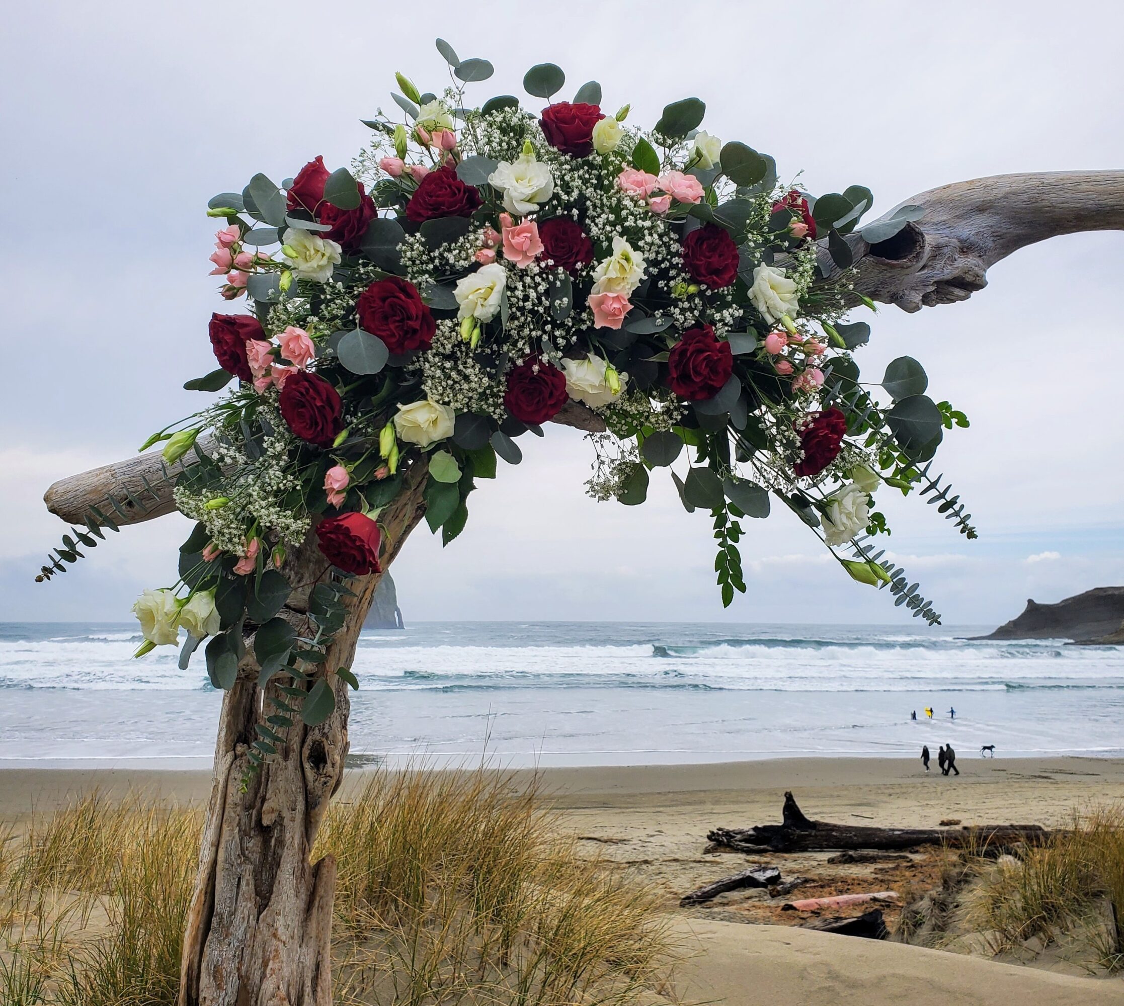 Large floral arrangement with red, pink, and white flowers on driftwood at beach, ocean and sky in background.