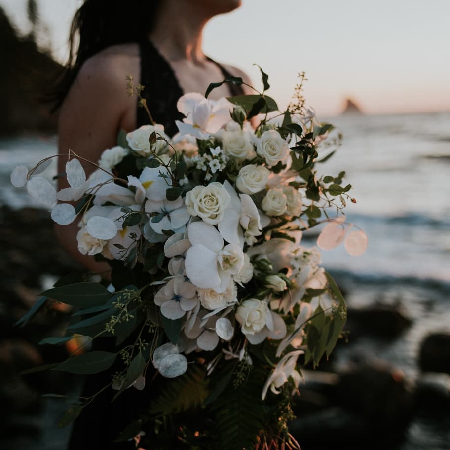 Person holding a large bouquet of white flowers near the ocean at sunset, with waves in the background.