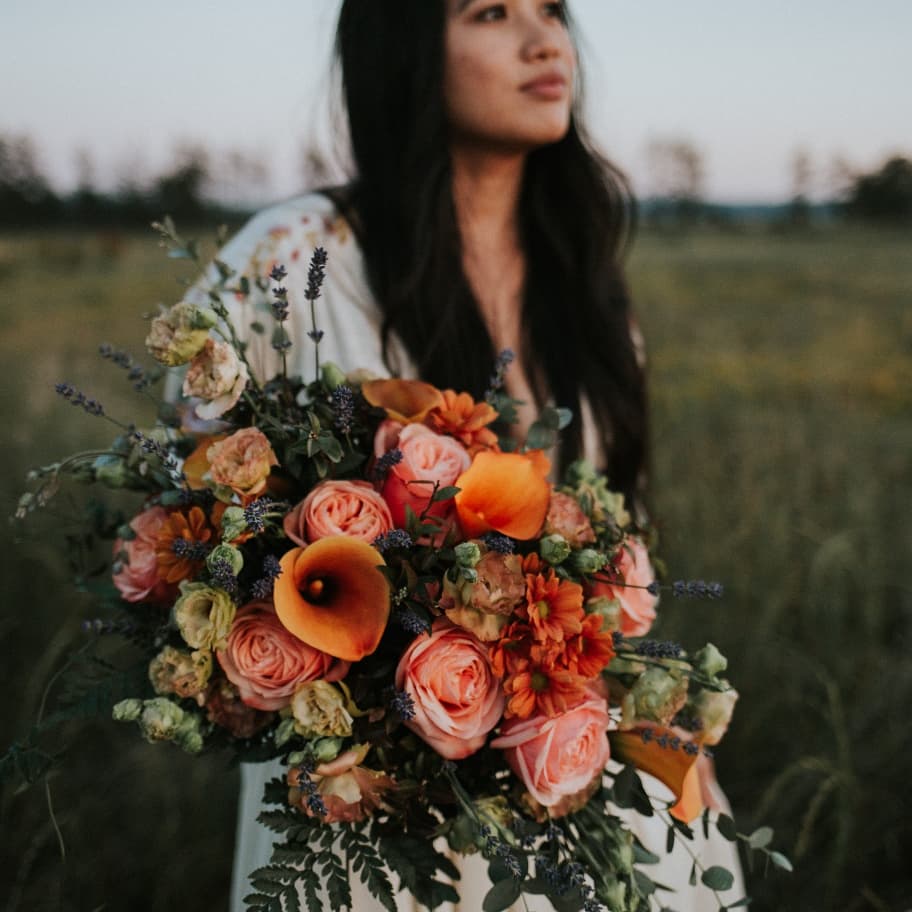Woman holding a large bouquet of orange, pink, and purple flowers outdoors, with a blurred natural background.
