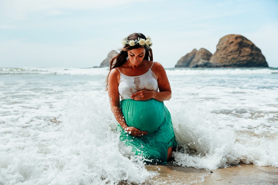 Pregnant woman in a white top and green skirt standing in ocean waves with rocks in background, wearing a flower crown.