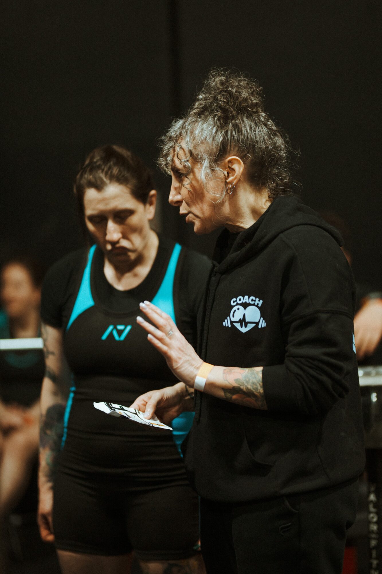 Two women in athletic clothing engaged in conversation, one holding a clipboard, in a dark indoor setting.