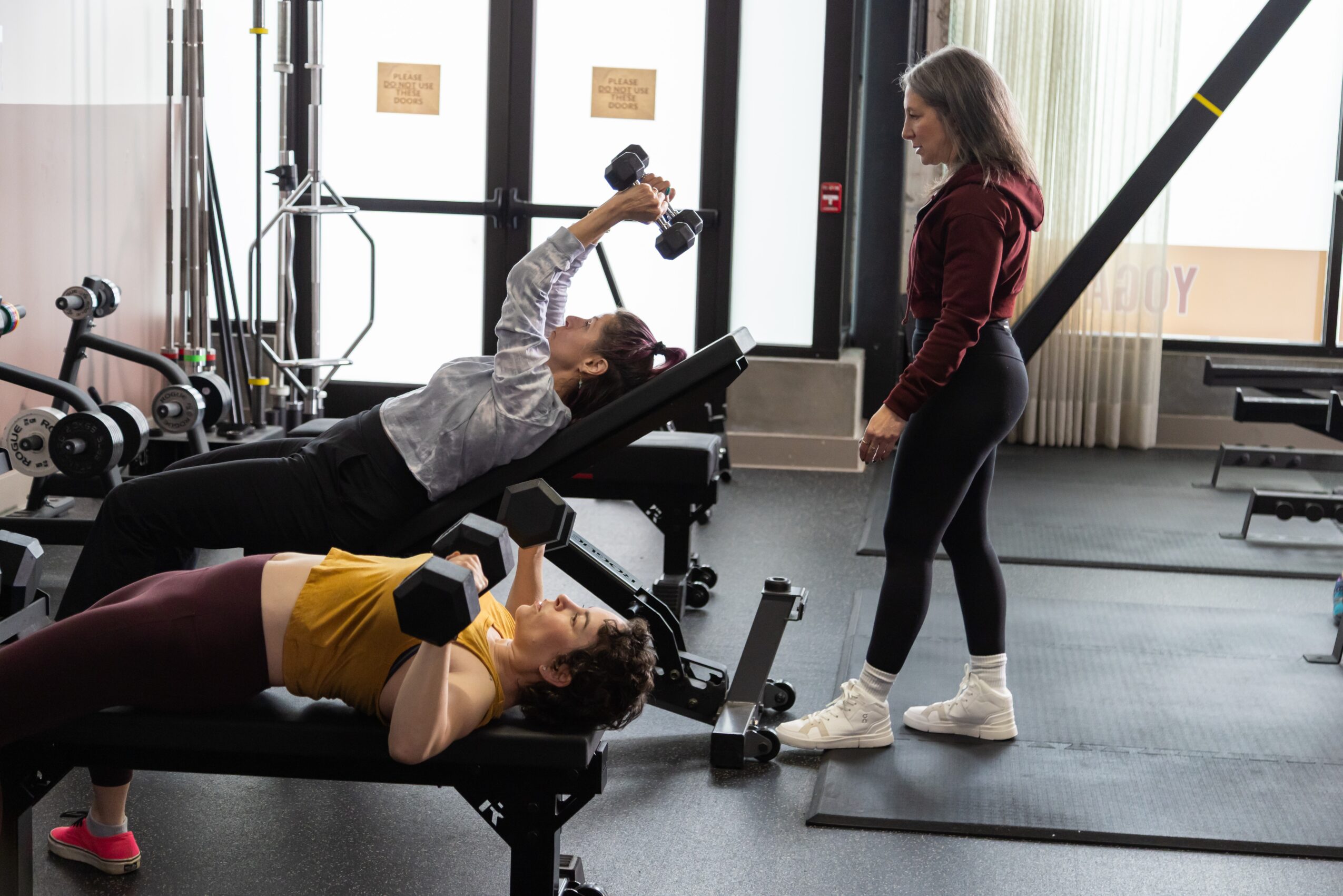 Two women exercise with dumbbells on workout benches, one standing and one lying down, in a gym with equipment and windows.