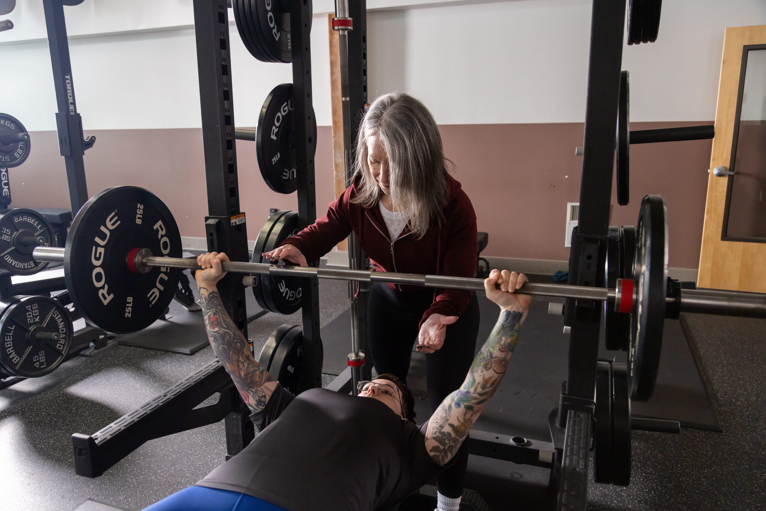 A woman with tattoos is lying on a bench, lifting a barbell with weights, while a young girl assists her.