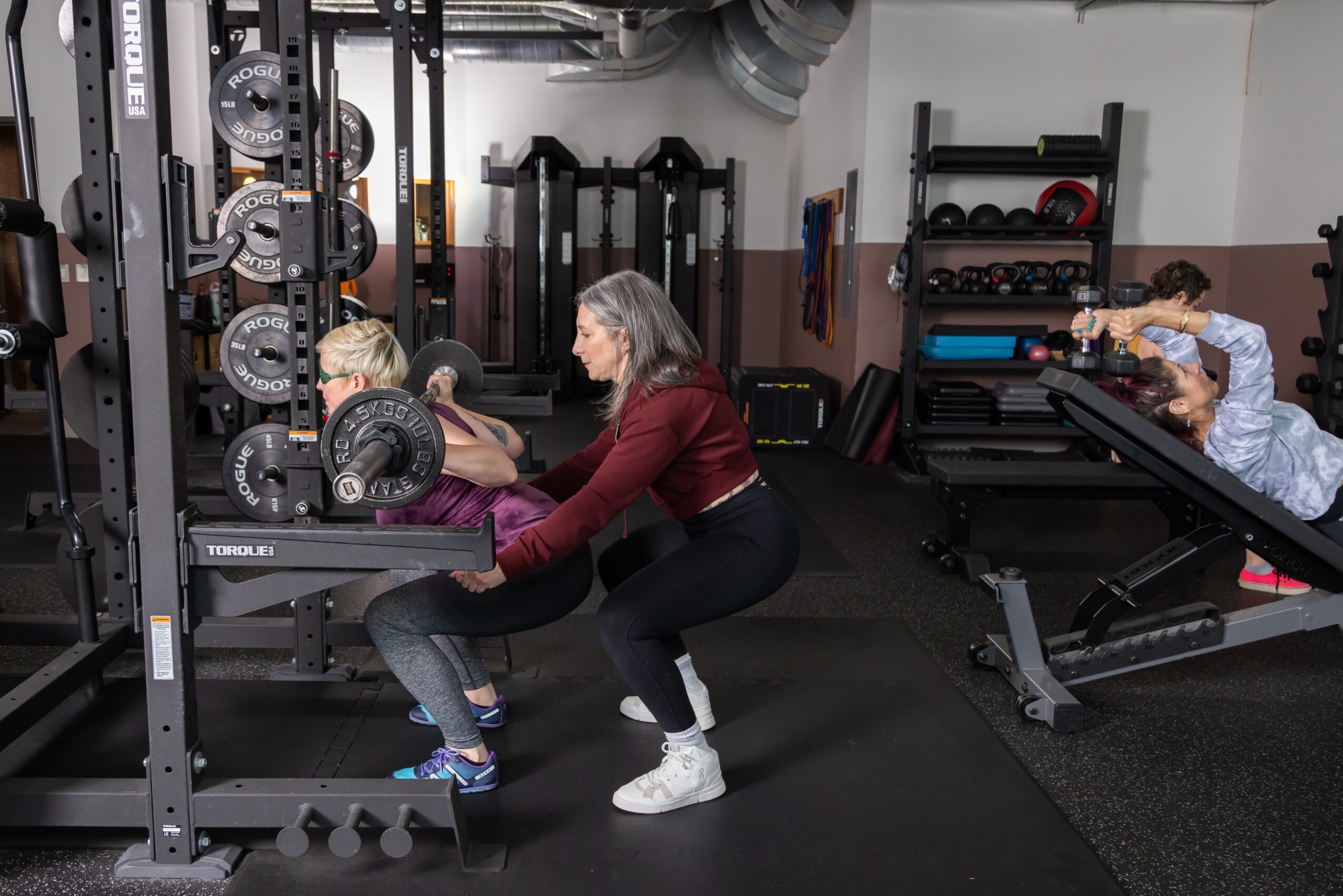 A woman and a girl are exercising with weights in a gym, with another girl on a bench nearby.