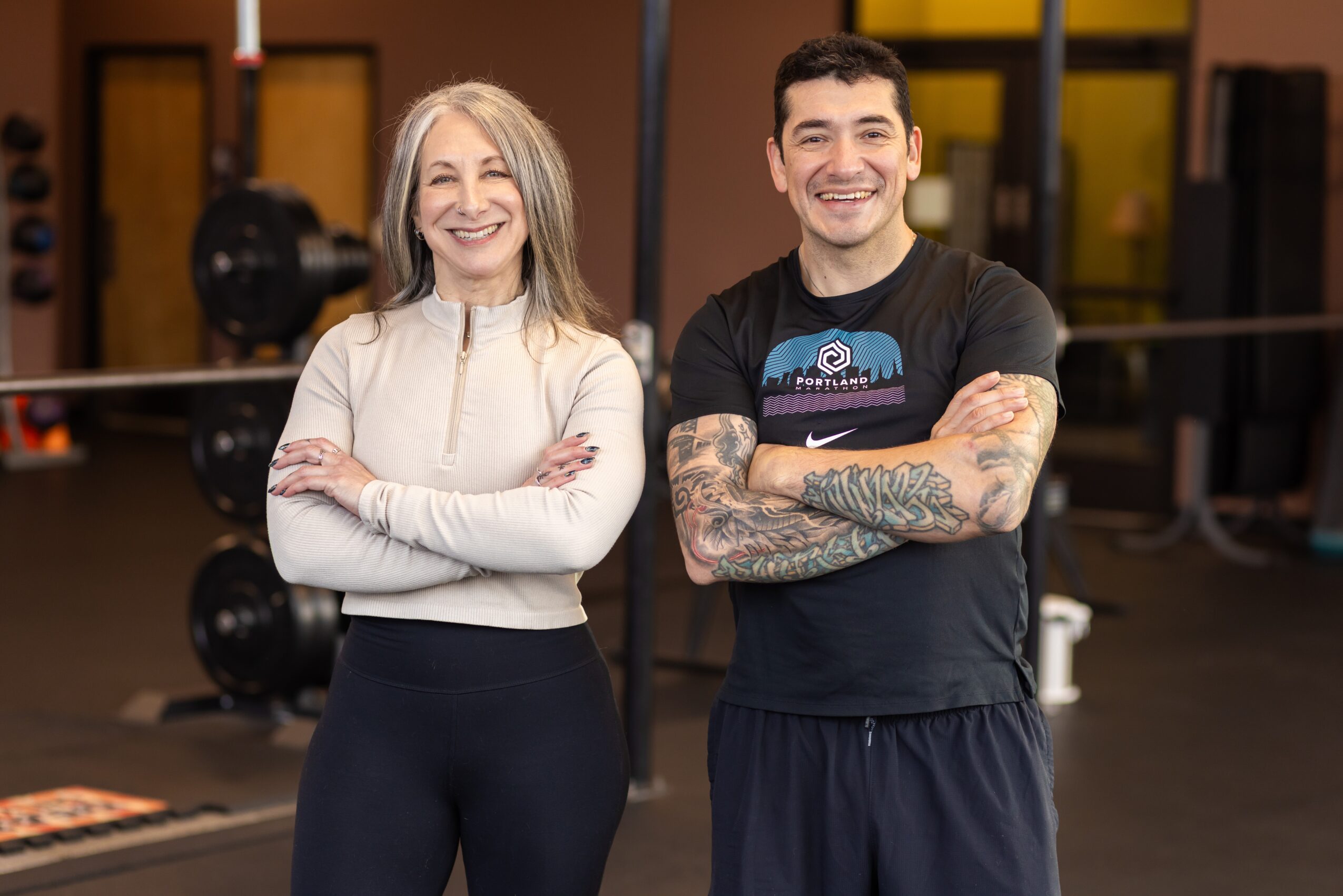 Two smiling people, a woman and a man, standing with arms crossed in a gym, smiling at the camera.