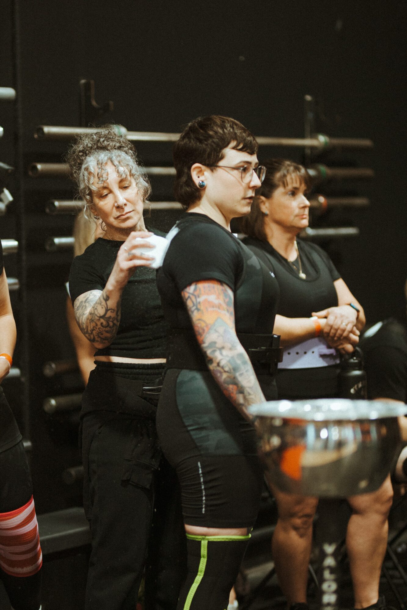 Three women standing in a dark room, one with tattoos, holding a glass and a bowl, with ballet barres in background.