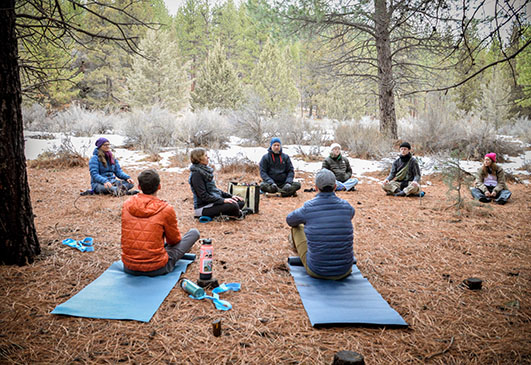 Group of people sitting on mats outdoors in a circle, surrounded by trees and nature, practicing meditation or yoga.