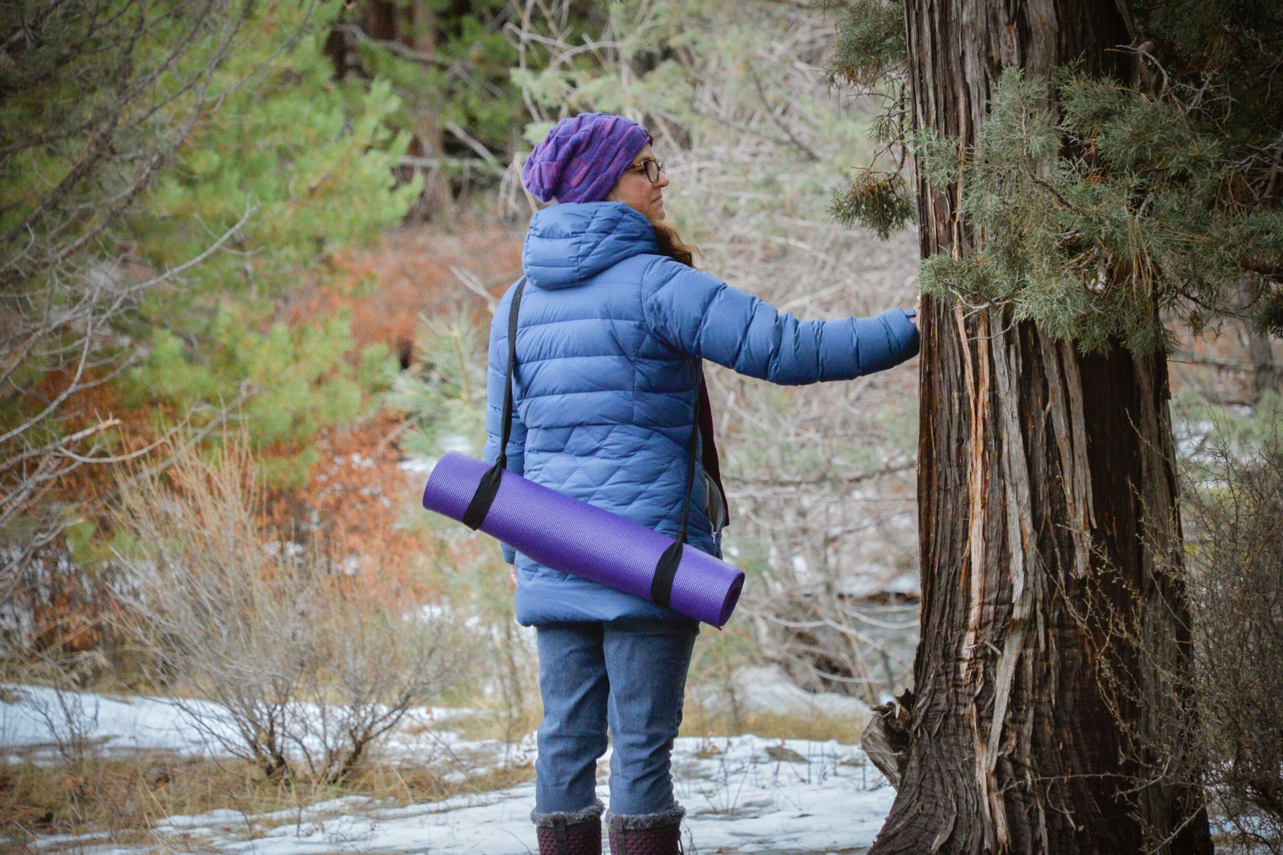 Person in blue jacket and purple headscarf touching a tree in a forested area with water and trees around.