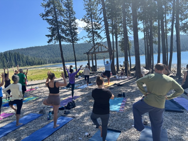 Group of people practicing yoga outdoors near a lake with trees and mountains in the background.