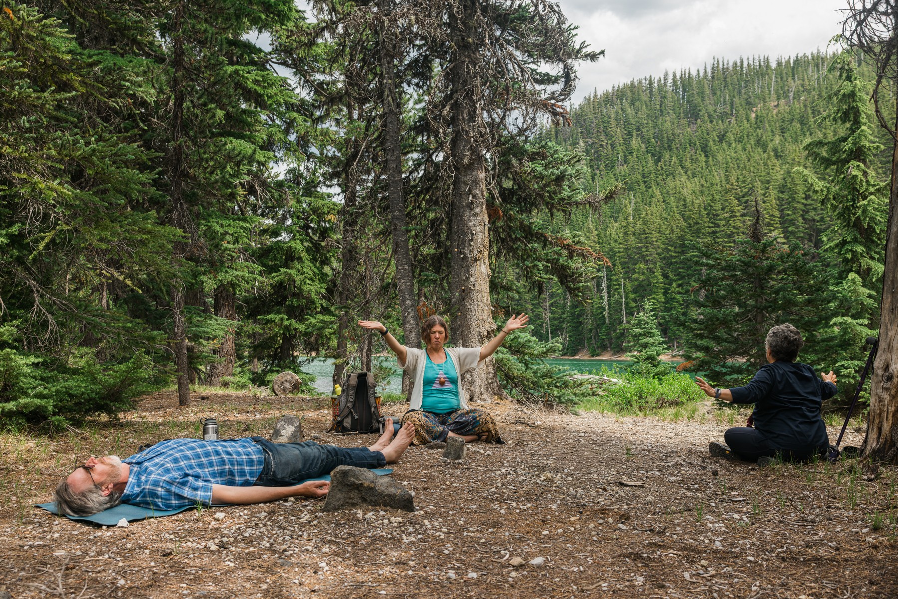 Three people in a forest clearing; one lying on the ground, one sitting cross-legged with arms raised, and one sitting on the ground facing away.
