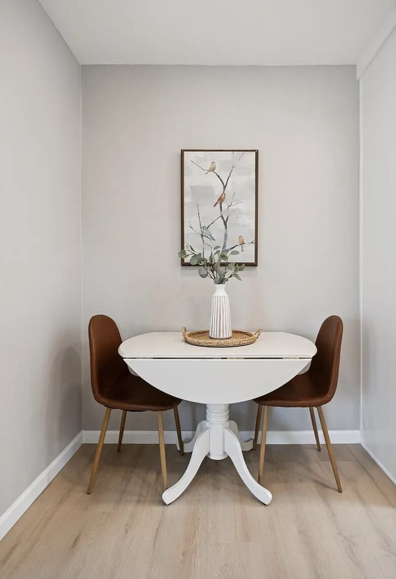 Small round table with two brown chairs, a white vase with flowers, and a framed artwork on a plain white wall.