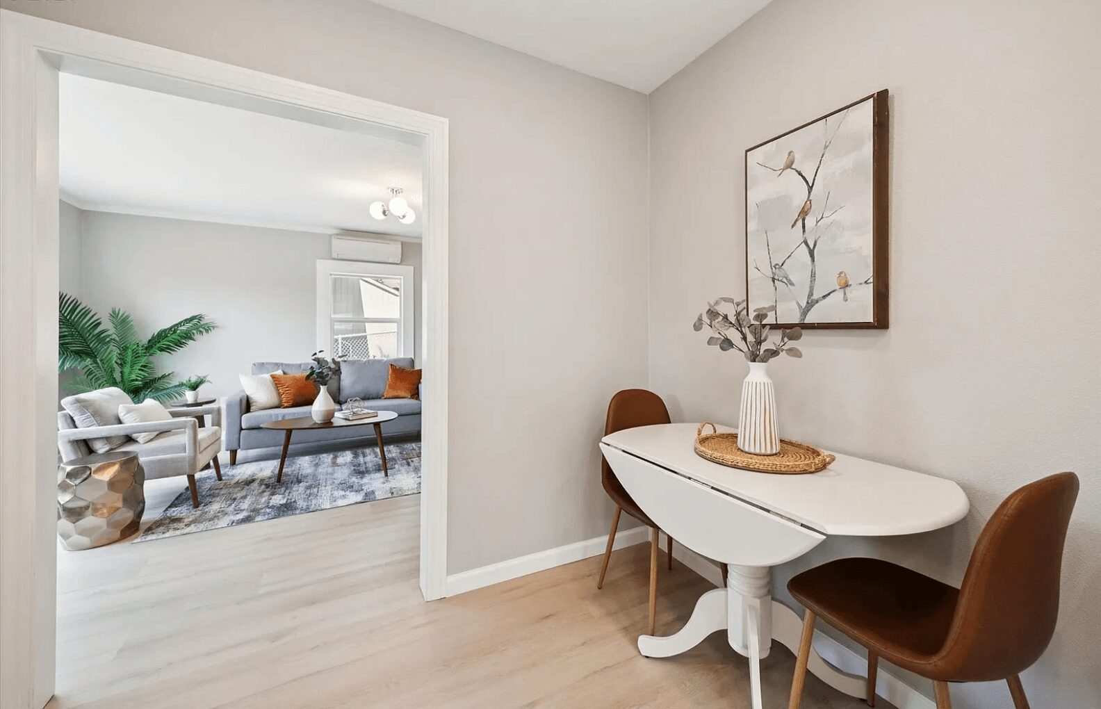 Corner of a room with a white table, two brown chairs, a vase with branches, and a framed artwork on the wall.
