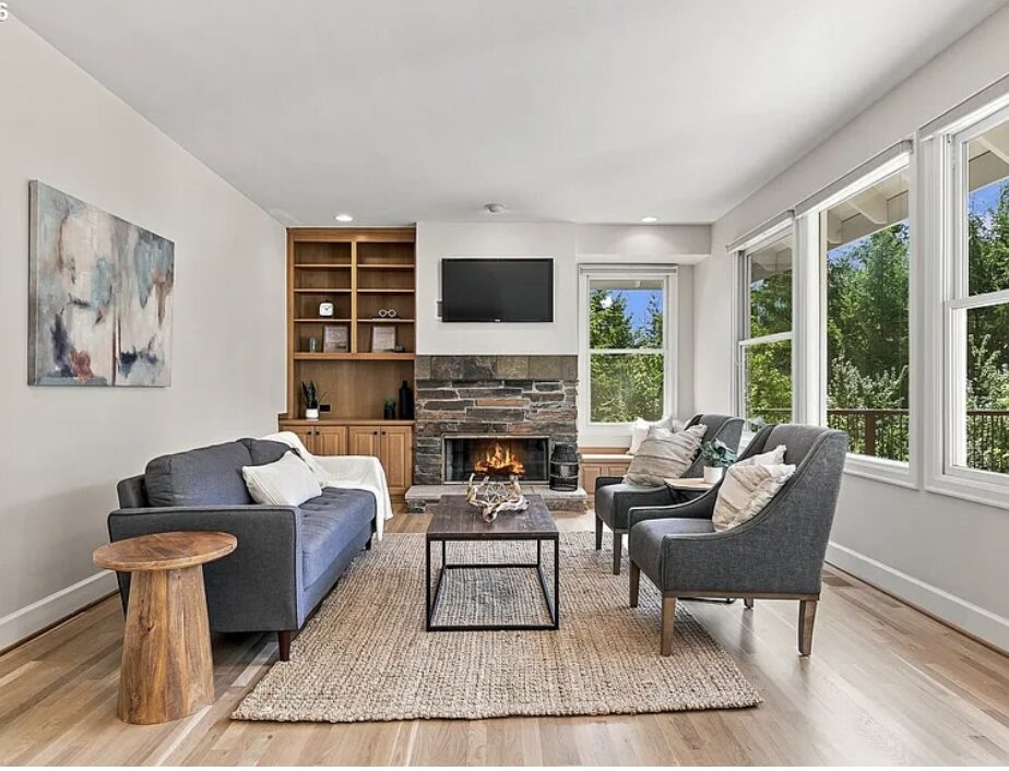 Living room with a sofa, two armchairs, a fireplace, a wall-mounted TV, large windows, and a wooden bookshelf.