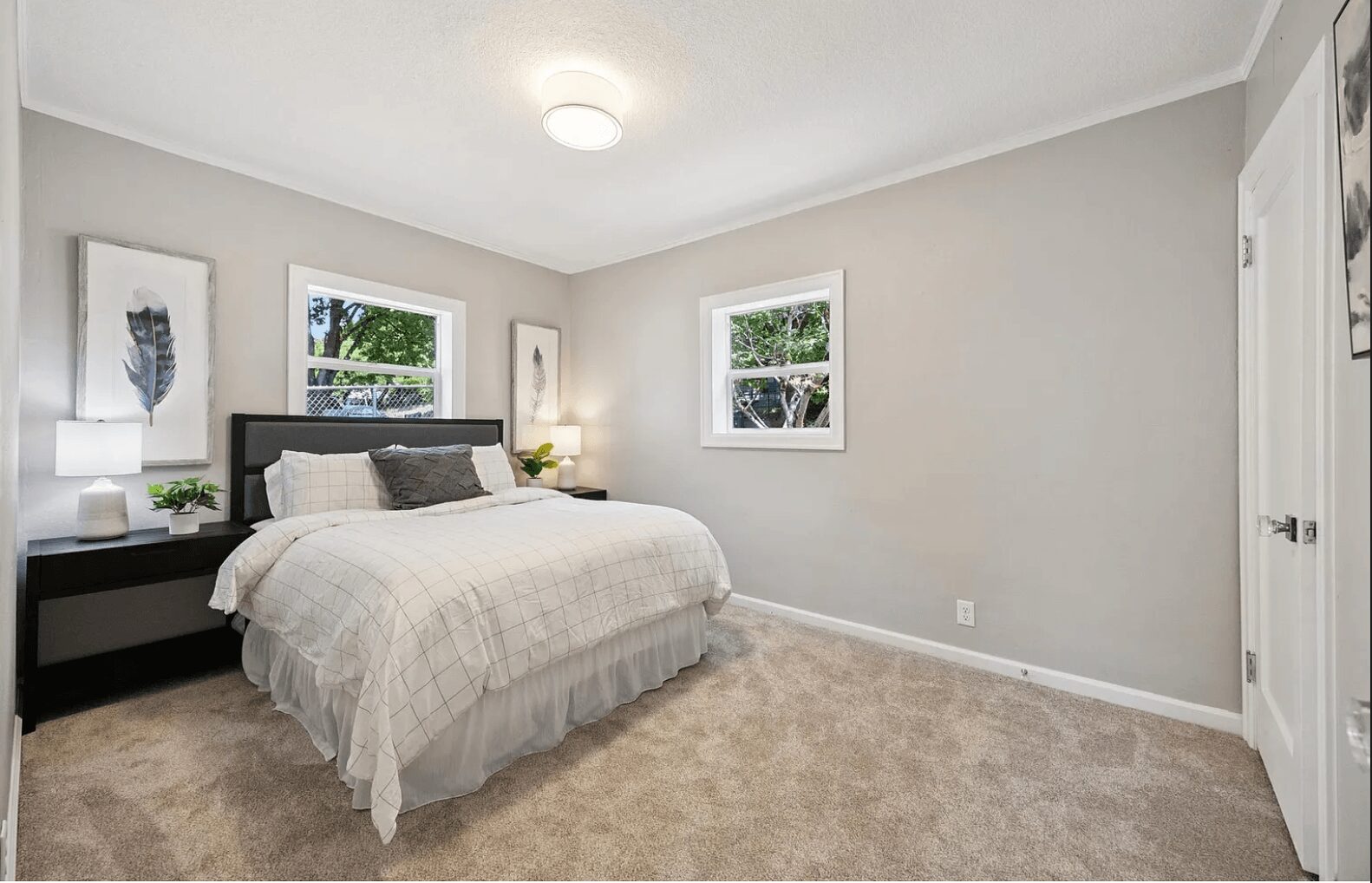 Bedroom with a bed, two windows, and wall art, neutral-colored walls, carpeted floor, and bedside lamps.