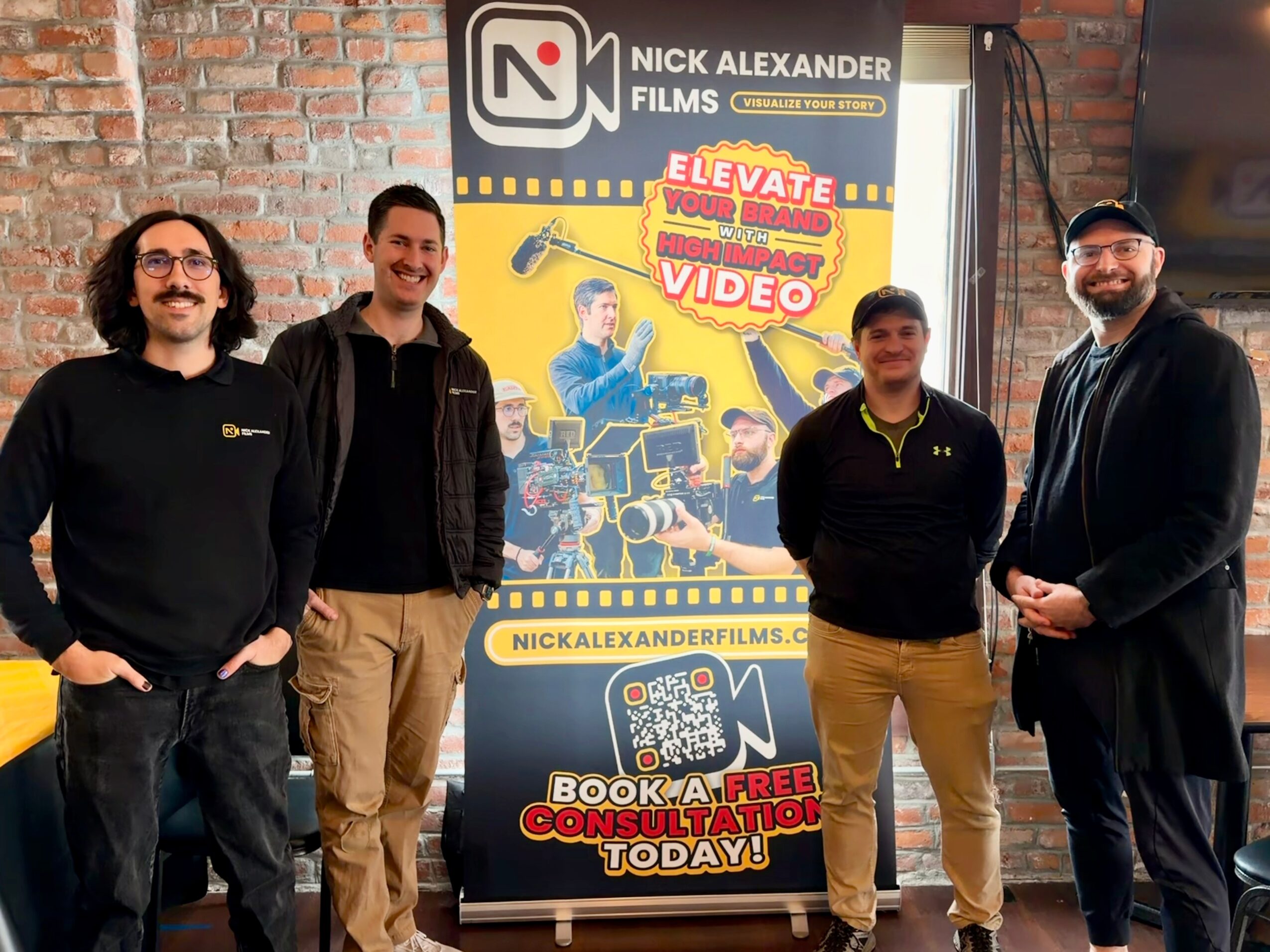 Four people standing in front of a Nick Alexander Films poster, smiling, with a brick wall background.