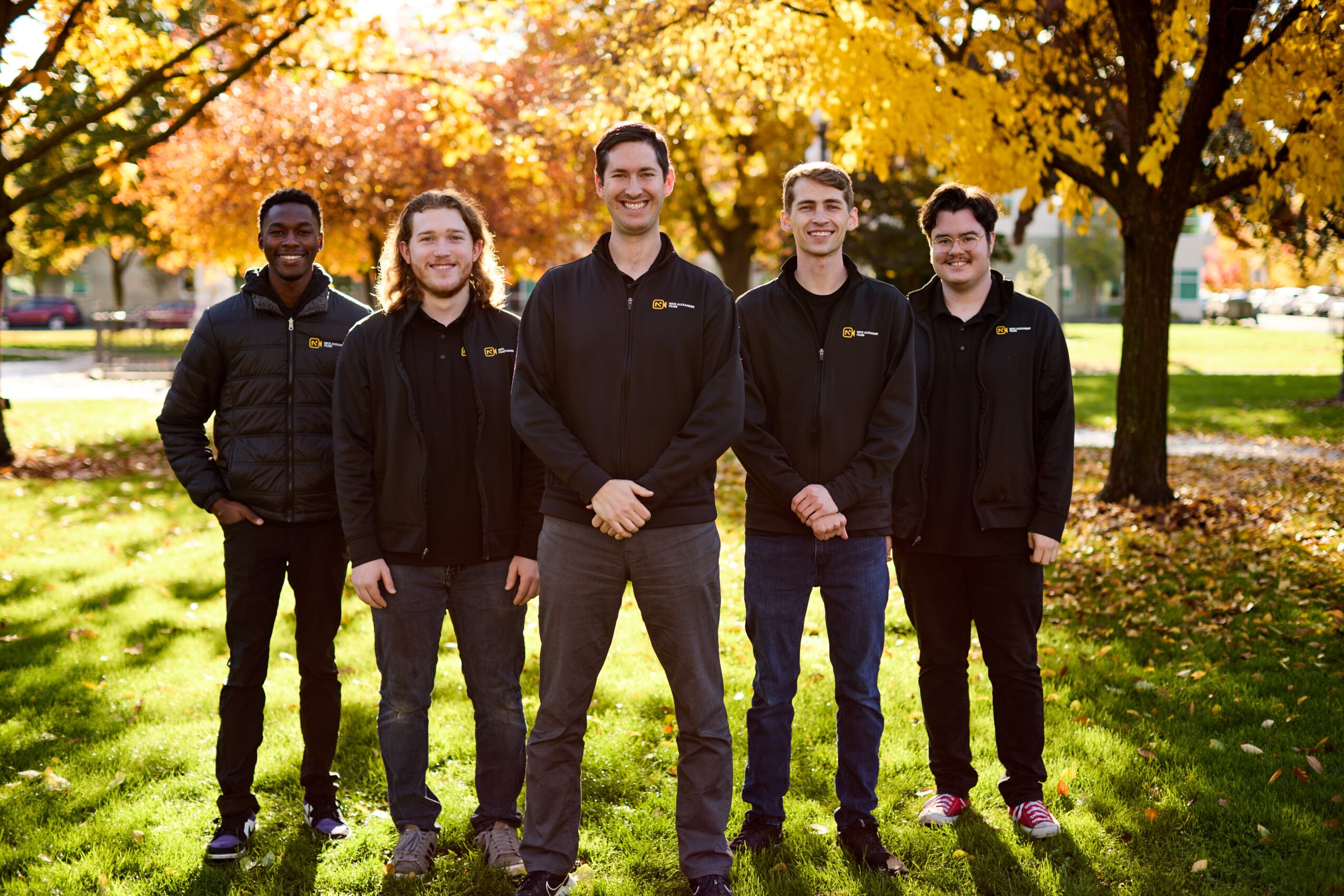 Group of five people standing outdoors in autumn with colorful trees and grass, smiling at the camera.