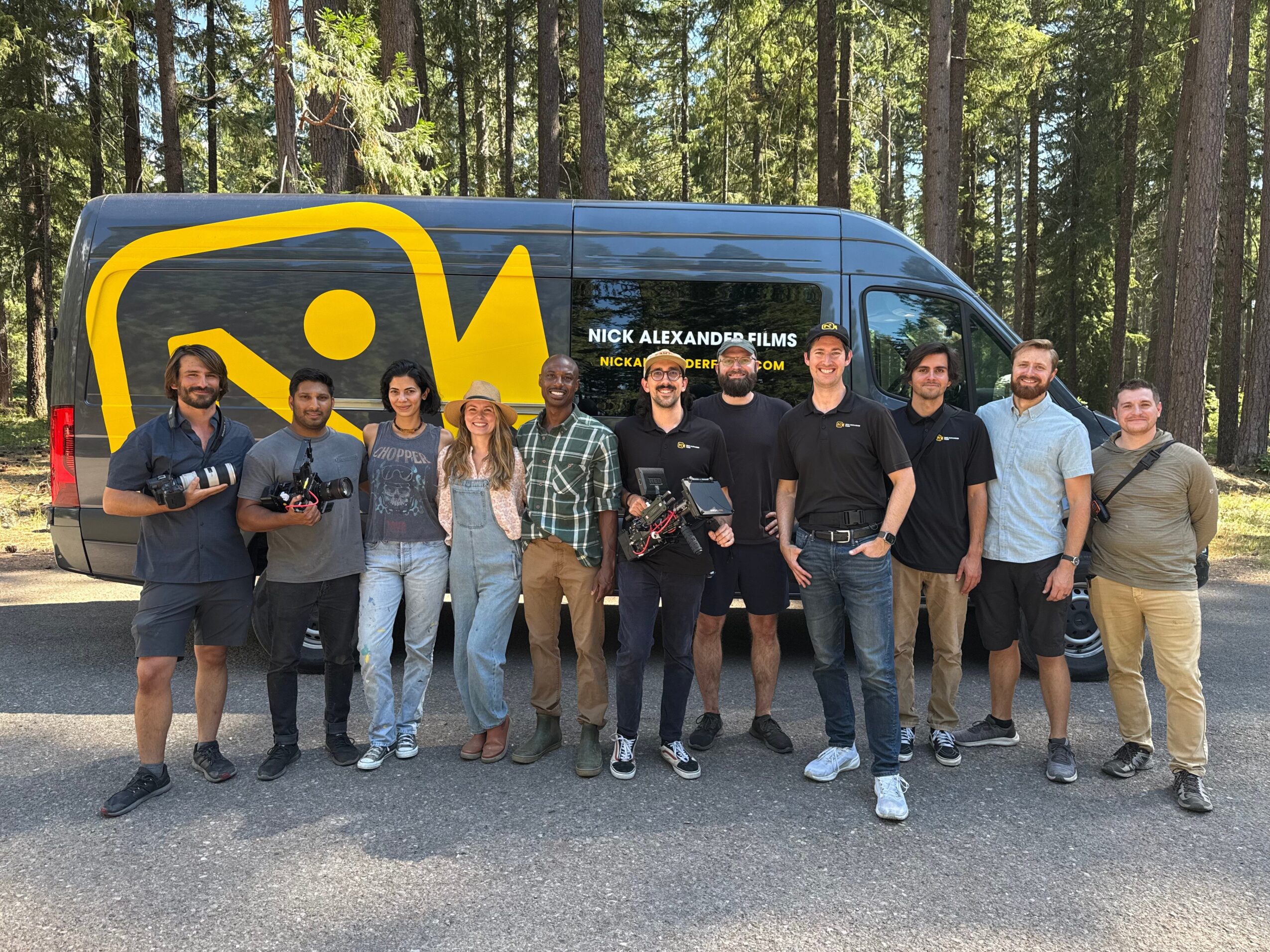 Group of ten people standing in front of a black van with yellow logo, in a forested area.