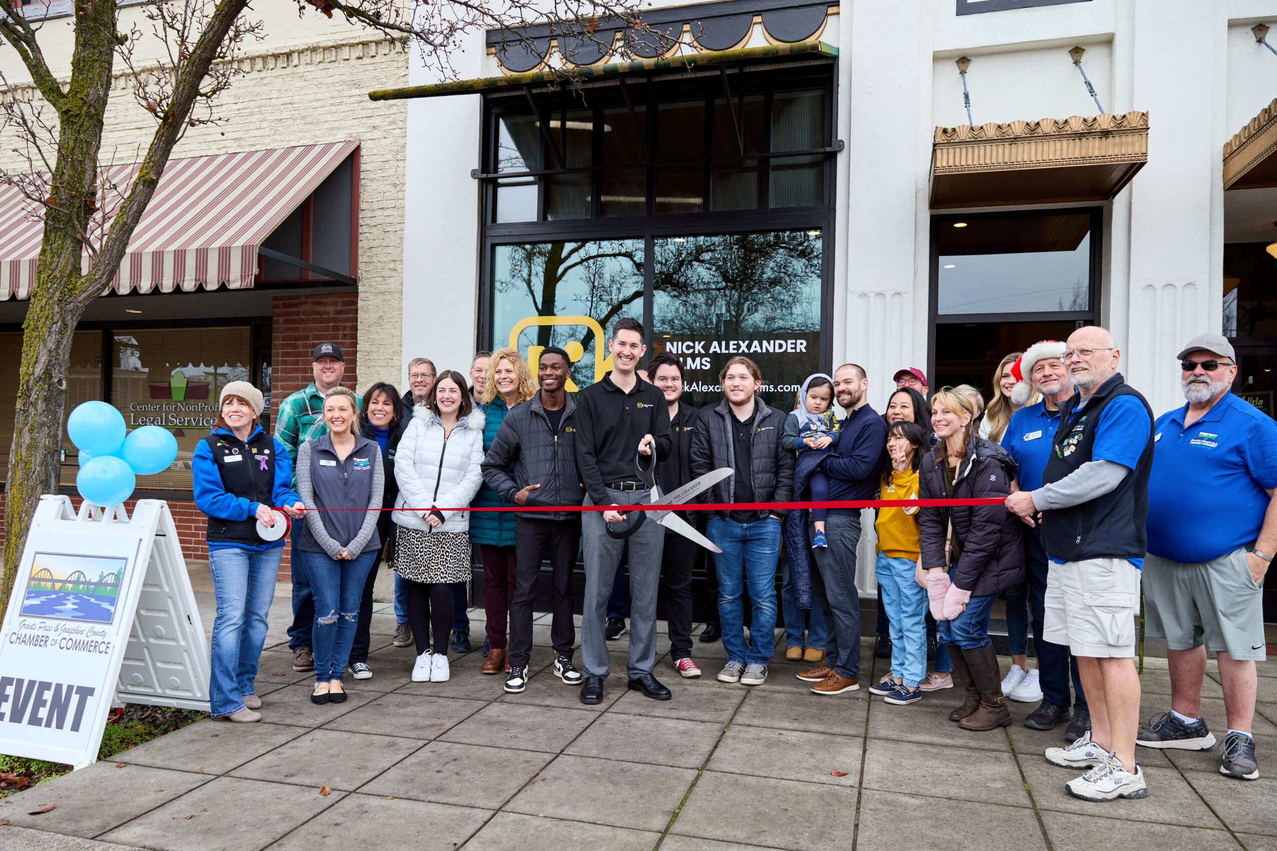 Group of people gathered outside a building for a ribbon-cutting event, with a red ribbon and scissors, some holding balloons.
