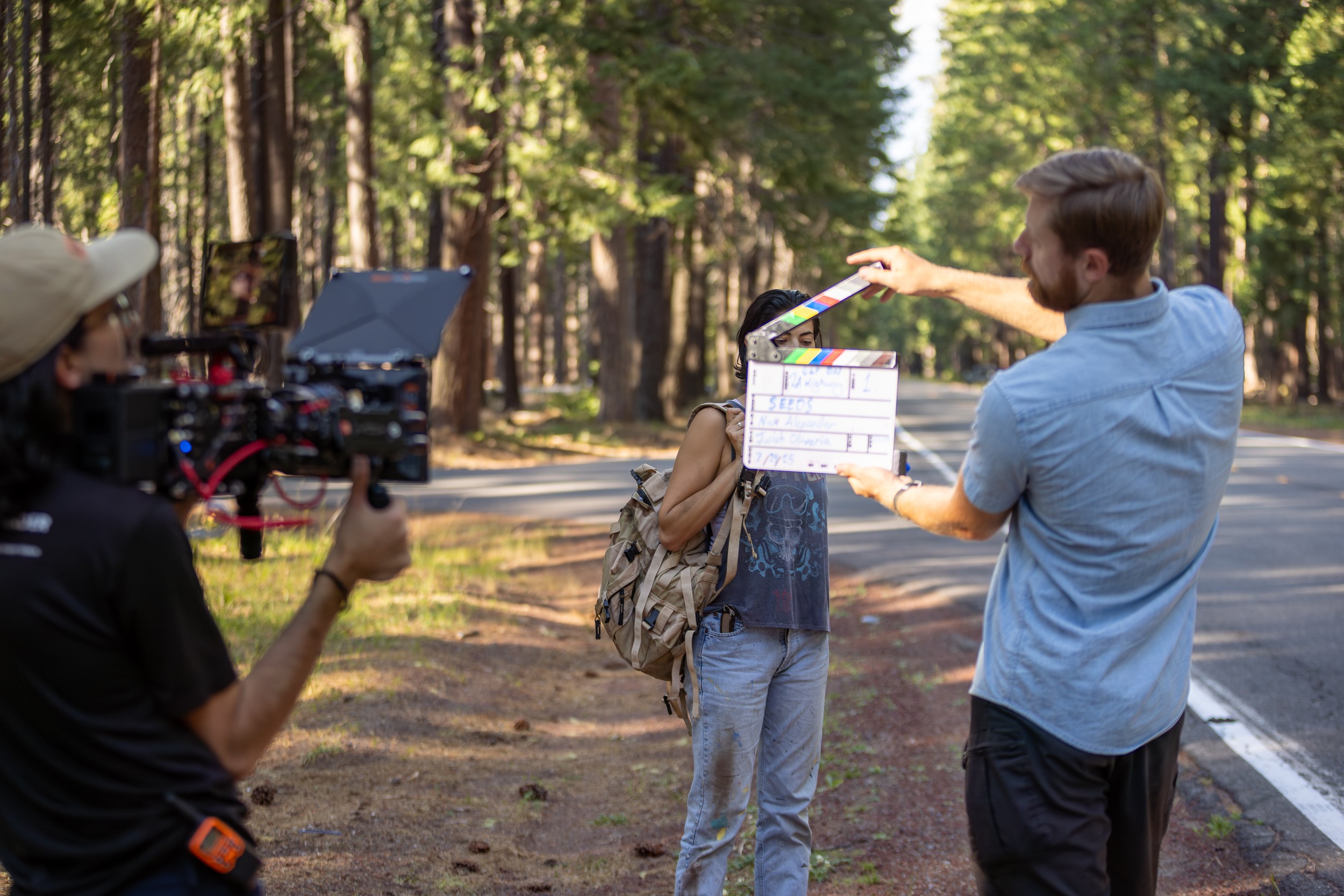 Filmmaker holding a clapperboard in a forest, directing a person with a backpack, camera operator filming nearby, trees in background.