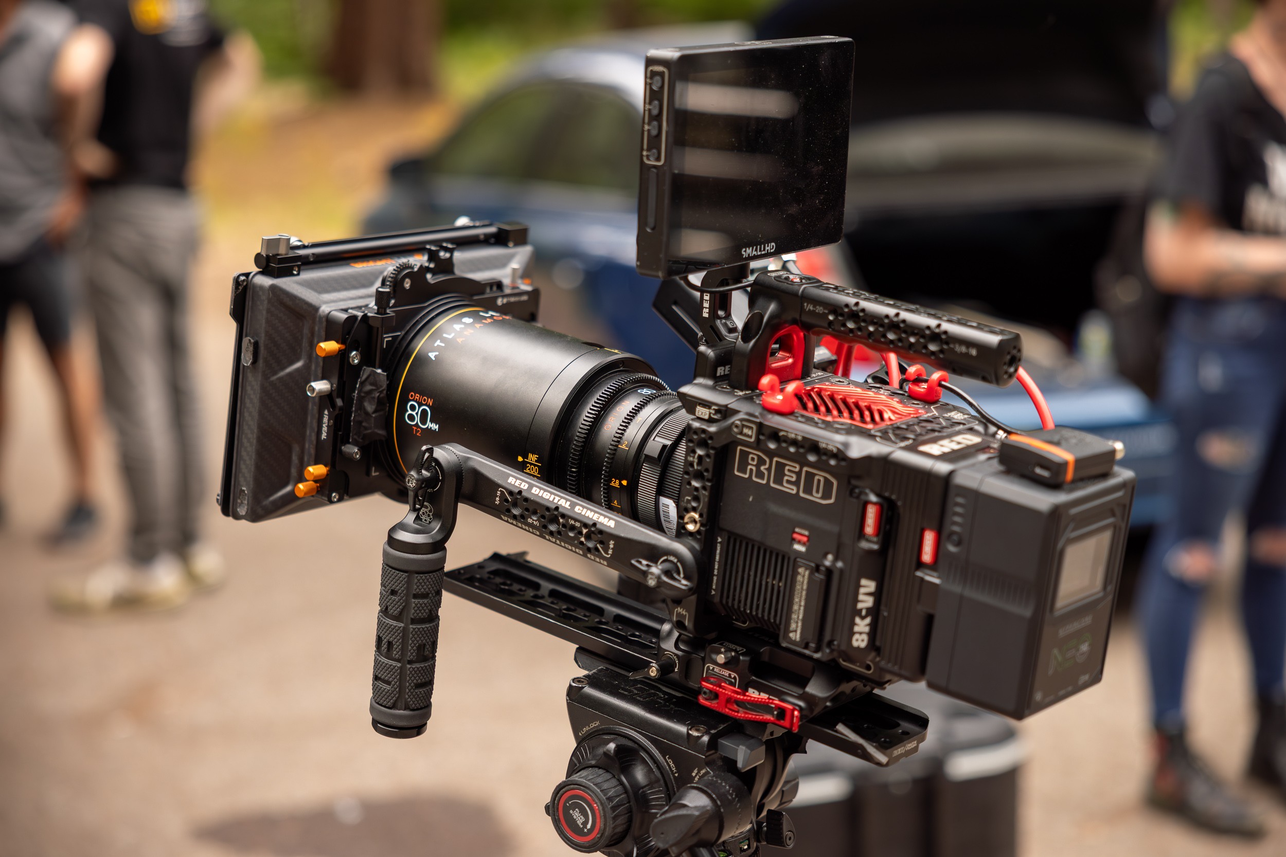 Professional camera mounted on a stabilizer rig outdoors with people and a vehicle in the background.