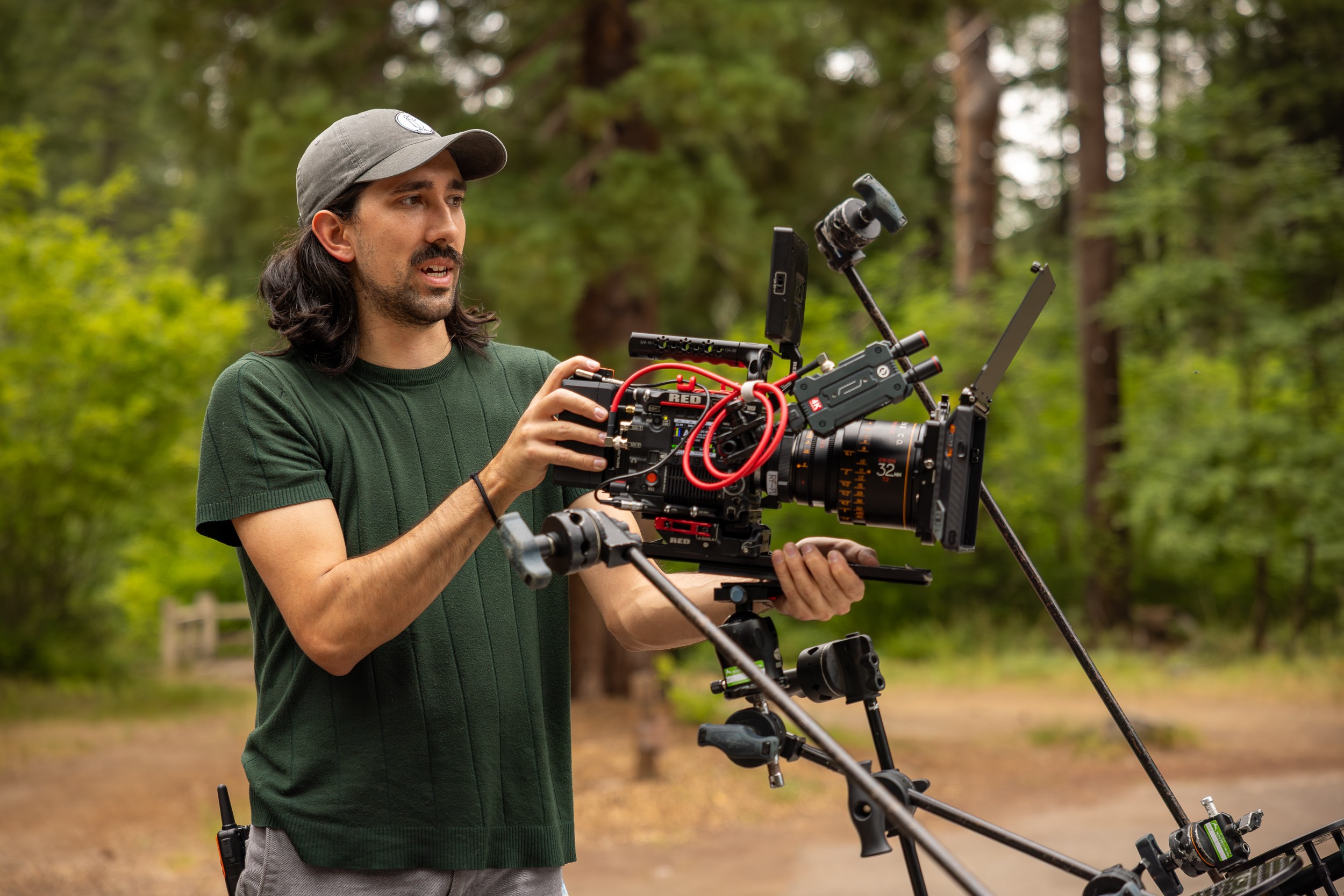 Man operating professional camera equipment outdoors in a forested area, wearing a cap and green shirt.