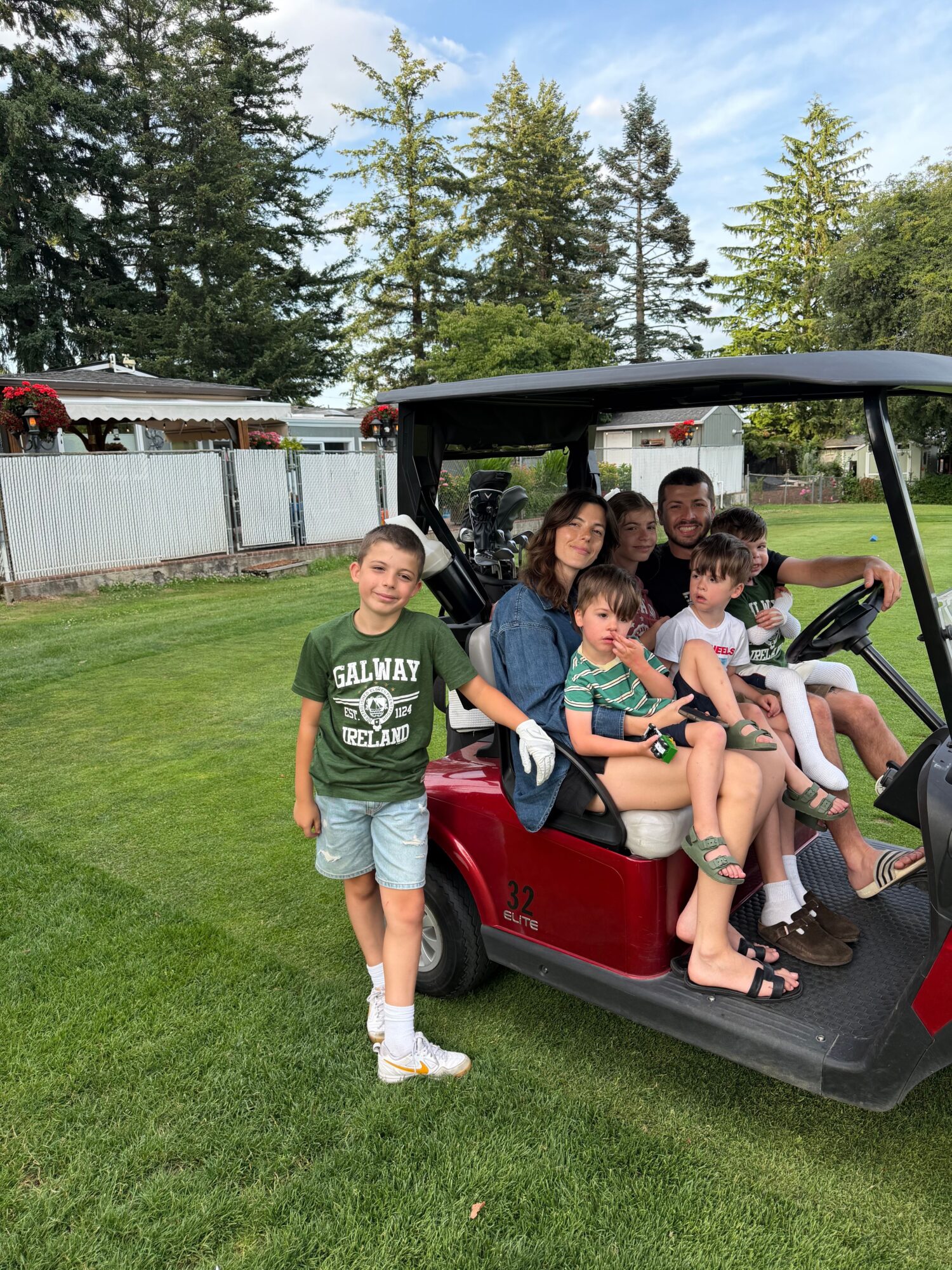 Four children sitting in a golf cart on a grassy area, with trees and a fence in the background.