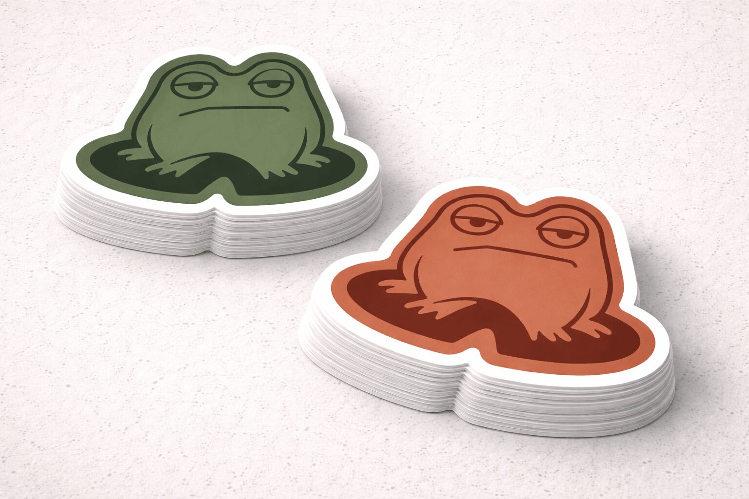 Stack of frog-shaped stickers, one green and one brown, on a white surface.