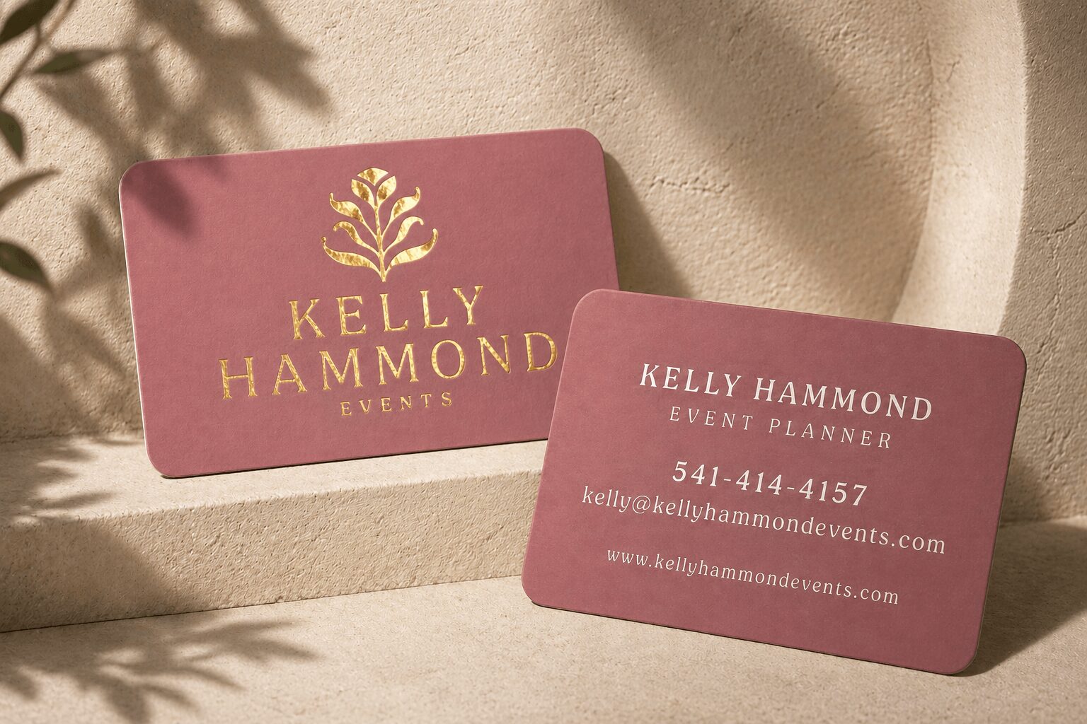 Two pink business cards with gold and white text, one displaying a logo and the other contact information, placed on a textured surface.