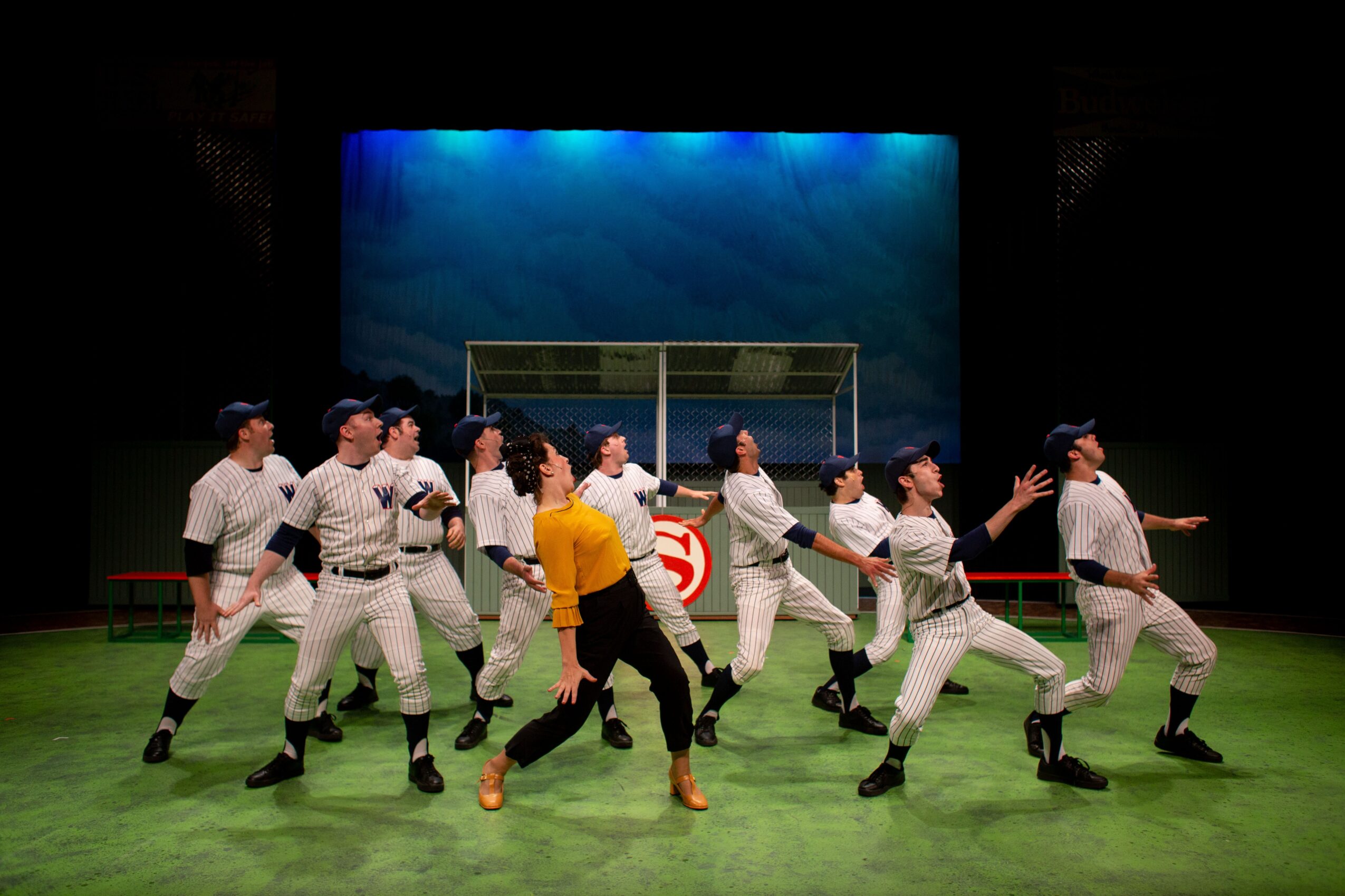 Theater stage with performers in baseball uniforms and a person in a yellow shirt, with a blue backdrop and green floor.