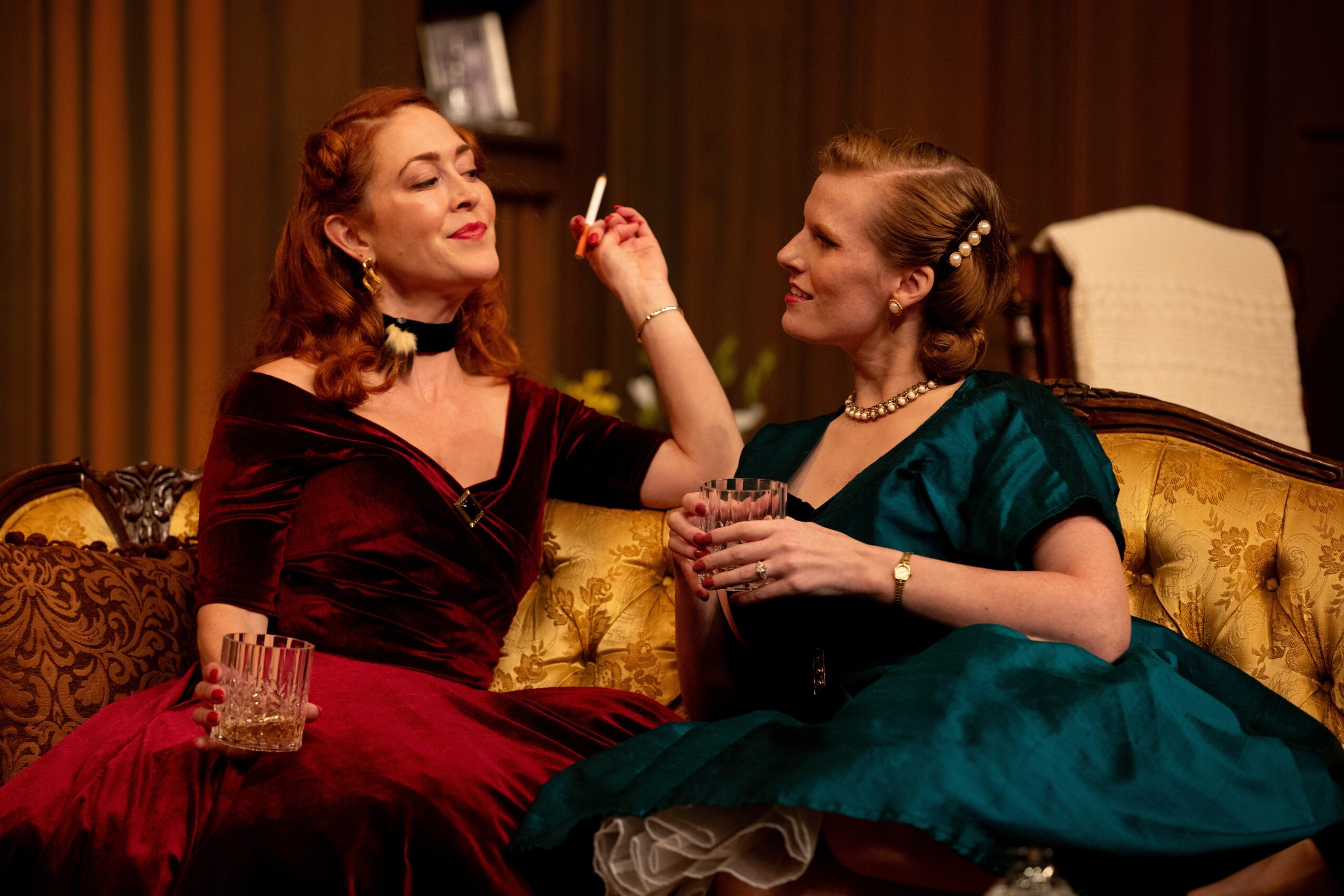 Two women in vintage dresses sit on a sofa, one holding a glass, the other with a cigarette, engaging in conversation.