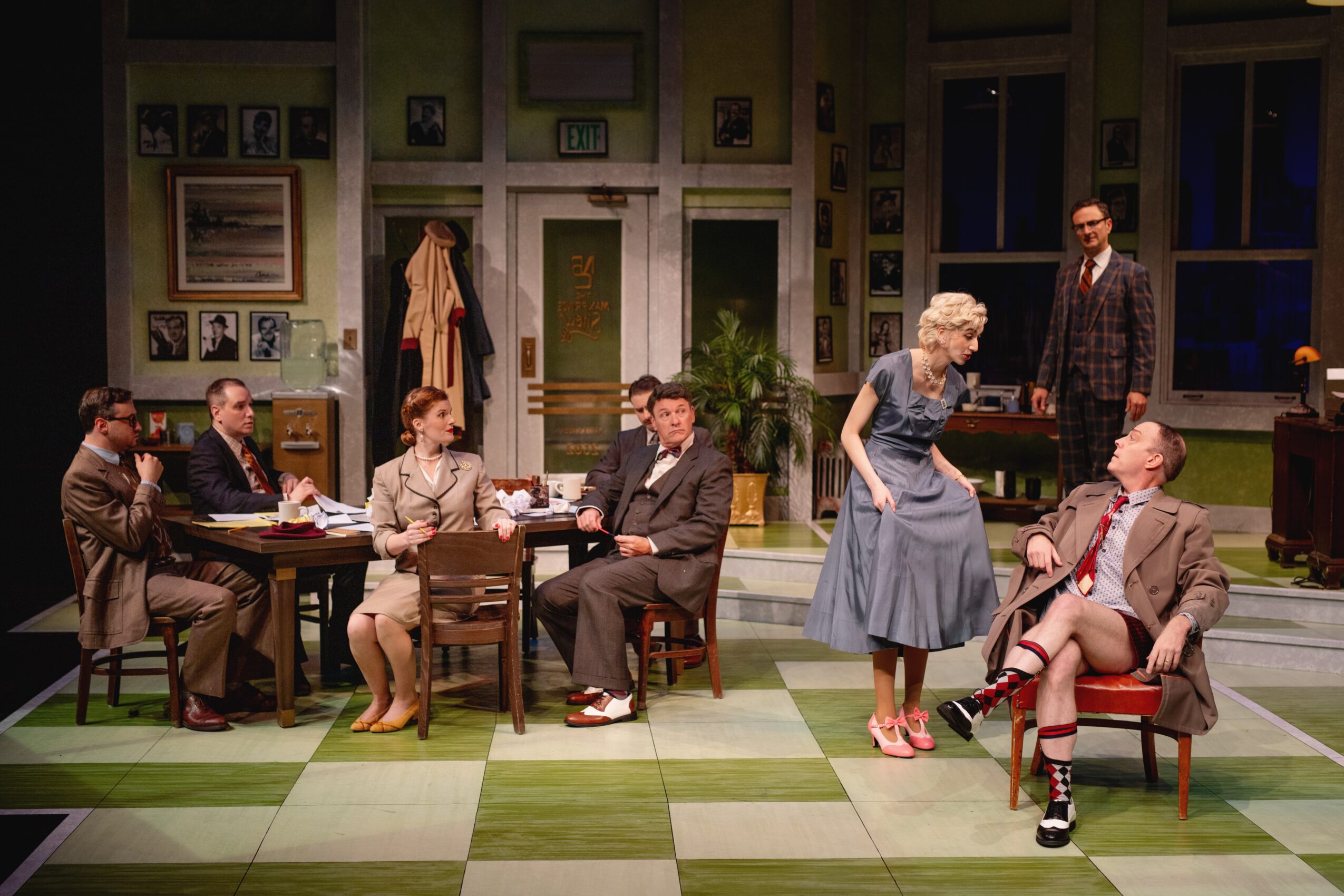 The scene shows a group of people in a room with green walls and large windows, some seated at a table, others standing or sitting, engaged in conversation.