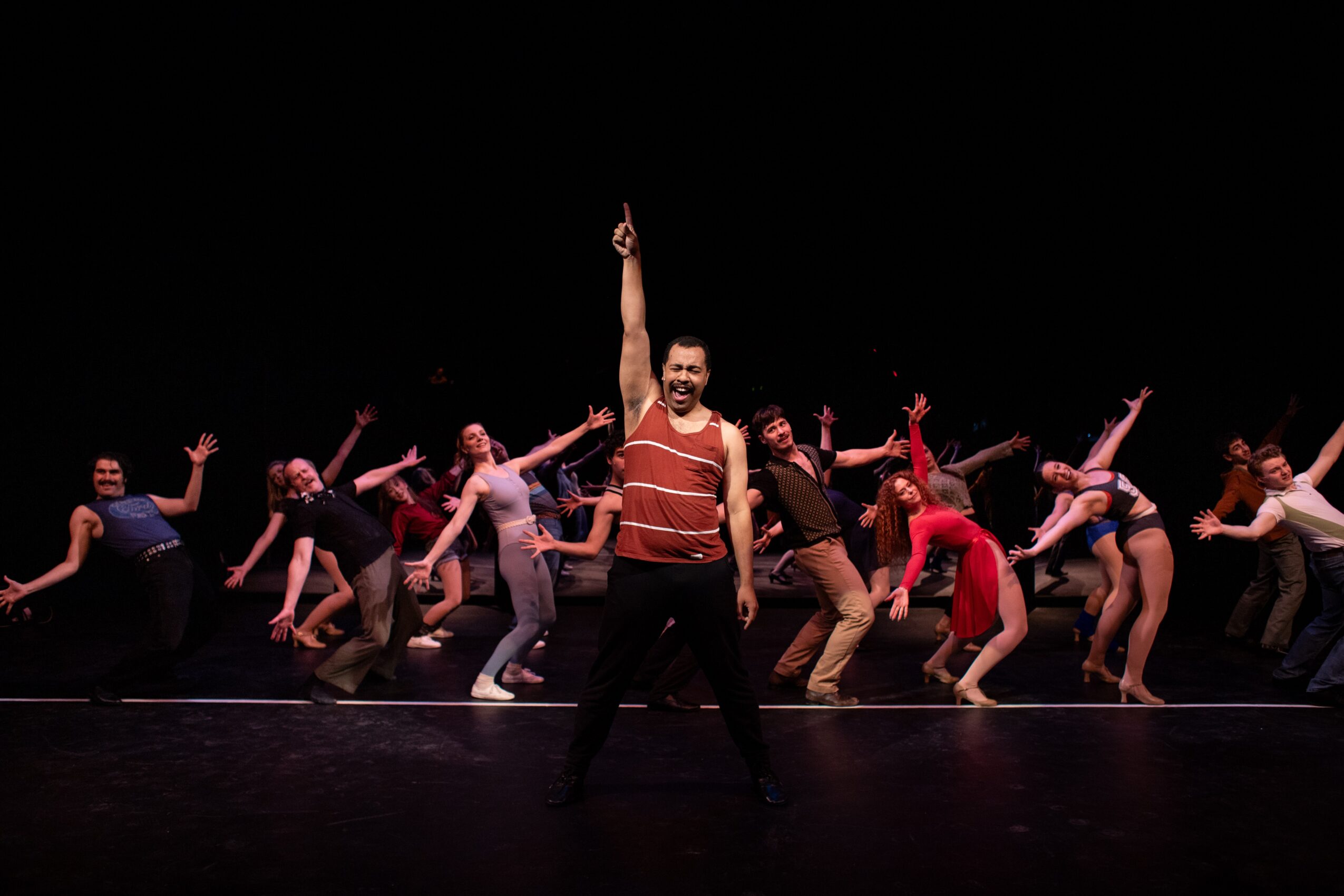 Group of dancers on stage with a man in the center raising one arm, all in various poses, dark background.