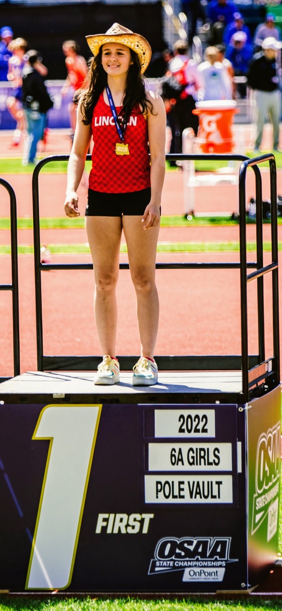 Young girl standing on a podium at a track event, wearing a red shirt, shorts, and a straw hat, with a crowd in background.