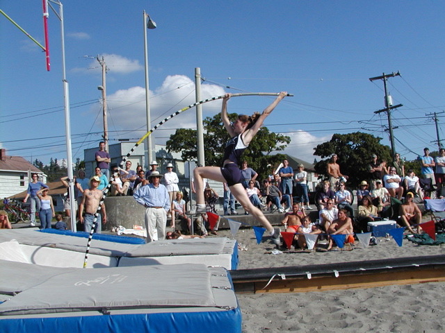 A person is mid-air jumping over a pole at a sports event with a crowd watching, blue sky, and power lines in background.