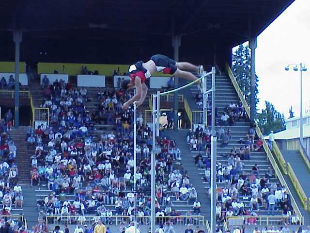 Athlete performing a pole vault over a bar at a stadium with spectators in the background.