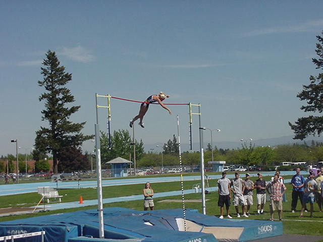 Person performing a high jump over a bar at an outdoor sports field with spectators and trees in the background.