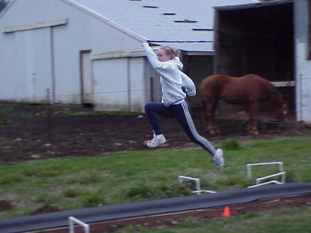 Girl jumping over a small obstacle outdoors with a horse in the background, holding a stick or pole, on grass and dirt.