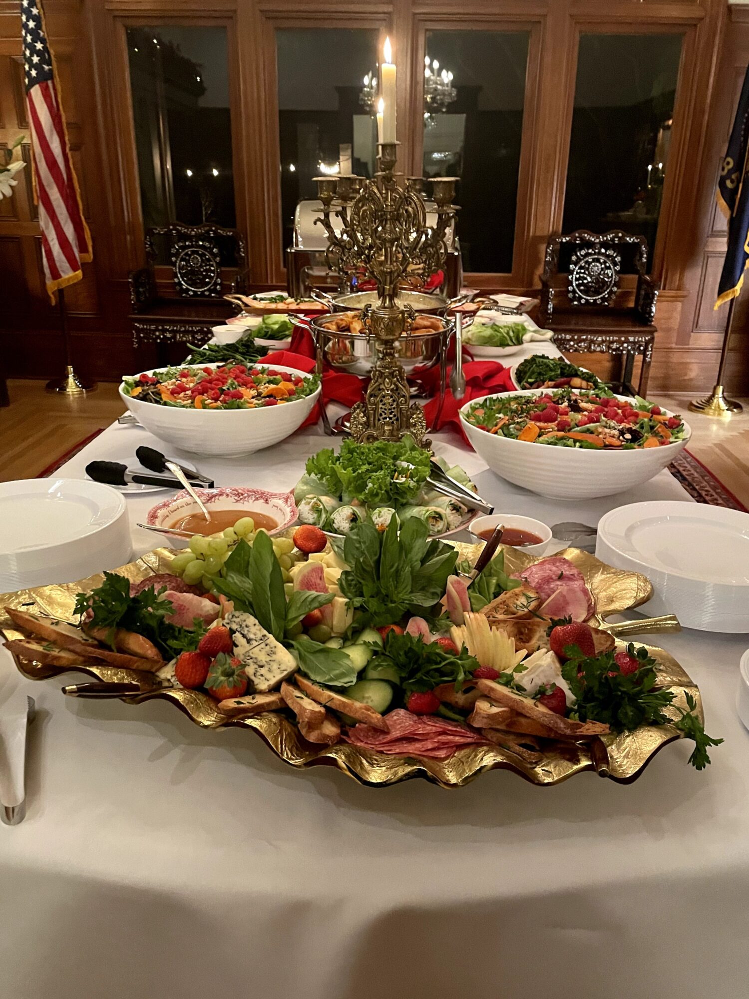 Table set with salads, a meat platter, and decorative items in a dining room with large windows and wooden furniture.