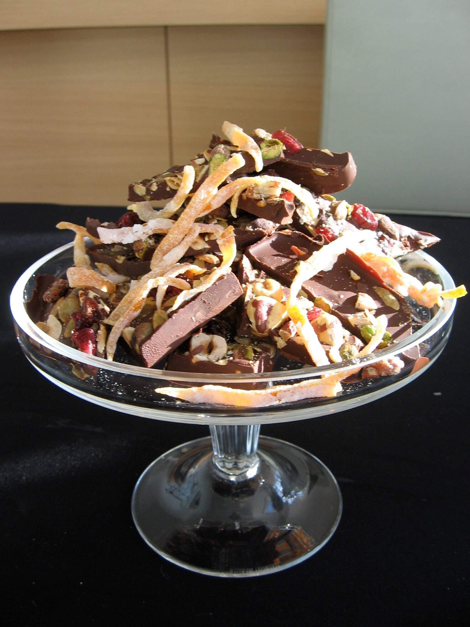 A glass dish filled with assorted chocolates and candies on a black surface, with a light-colored background.
