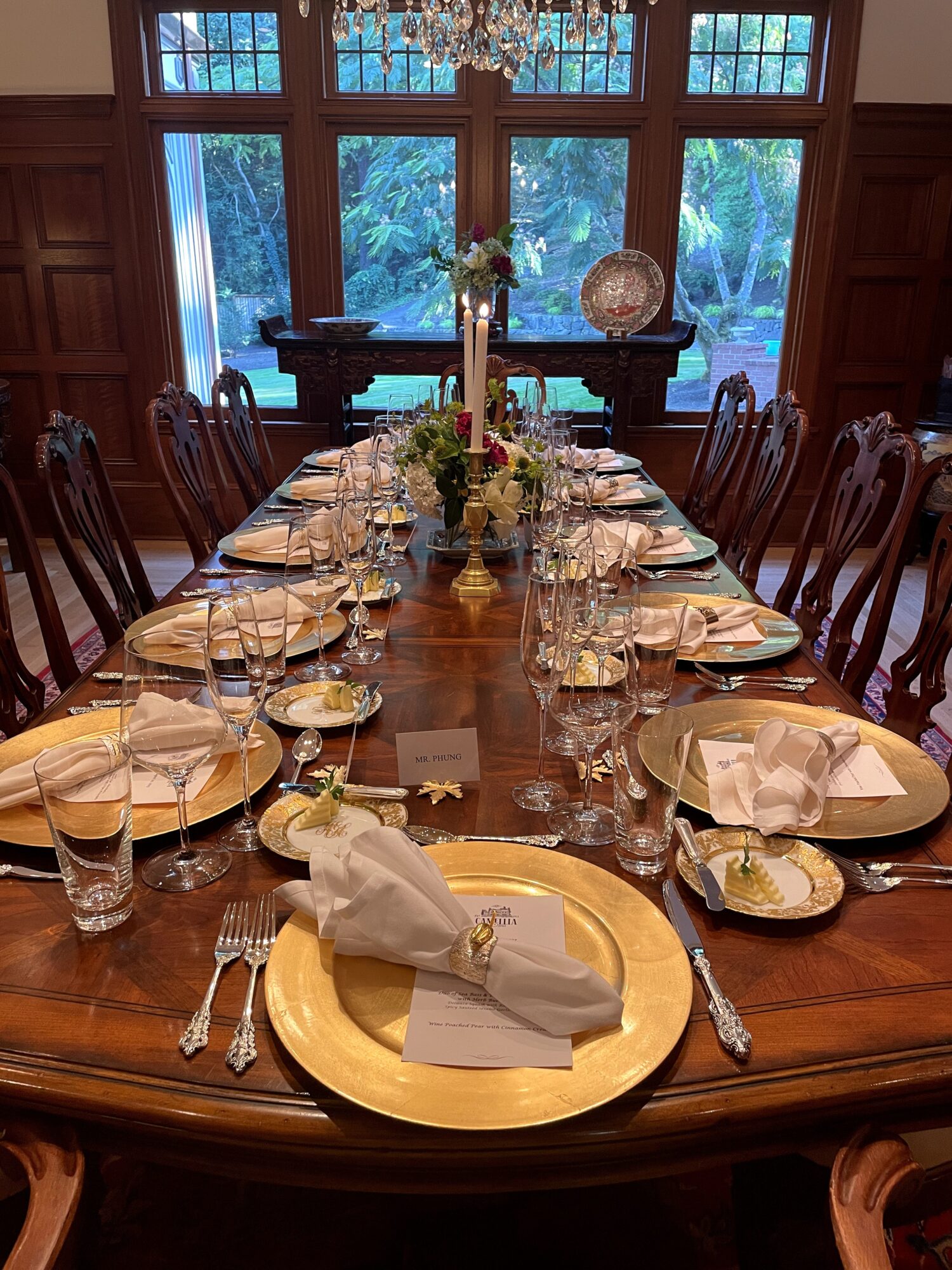 Long wooden dining table set with plates, glasses, napkins, and silverware, in a room with large windows and greenery outside.