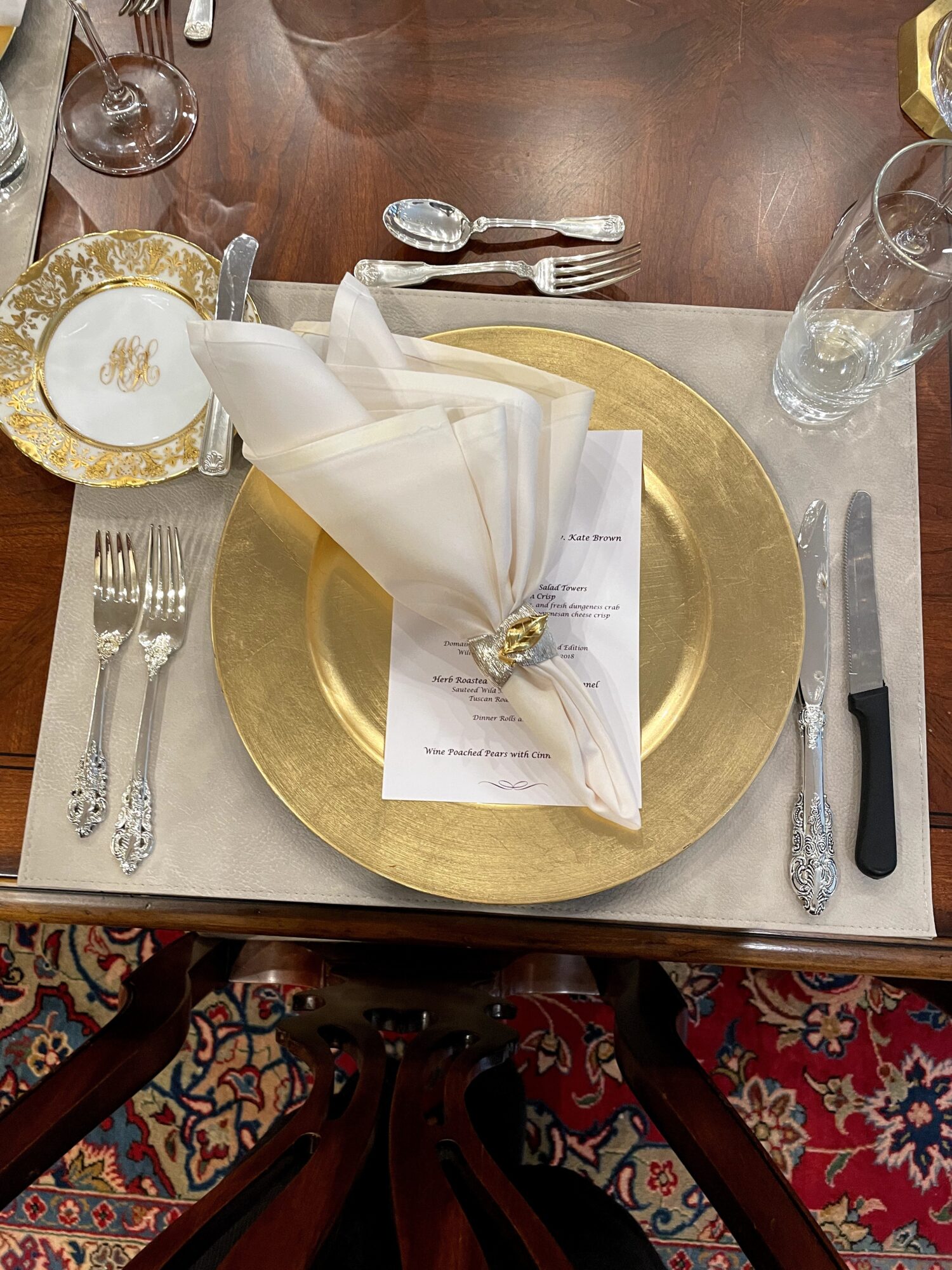 Table setting with a gold charger plate, white napkin, silverware, glass, and a menu, on a wooden table.