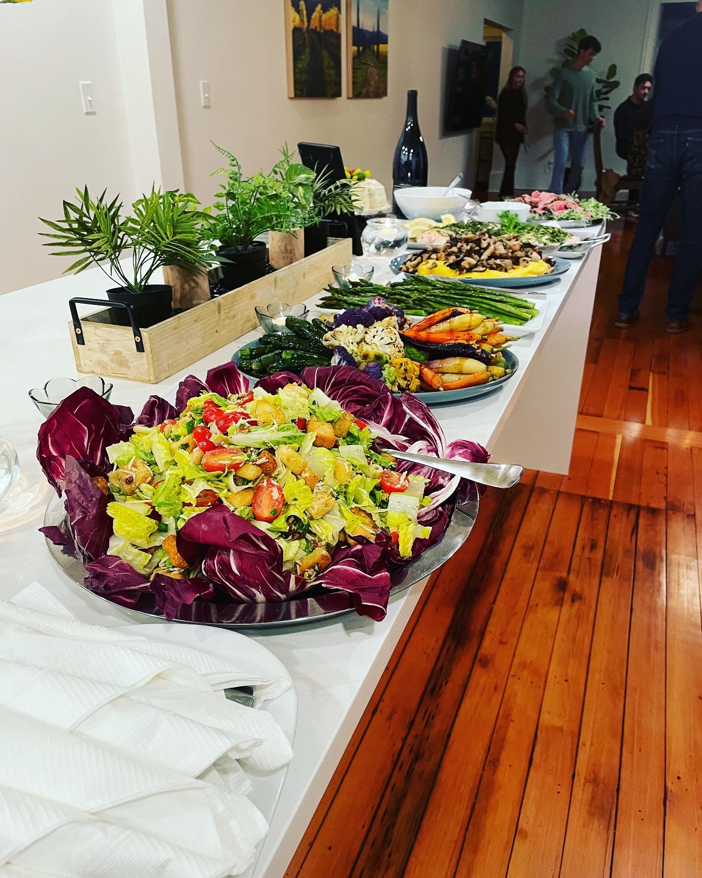 Buffet table with salads, grilled vegetables, and decorative plants in a room with wooden floor and people in the background.