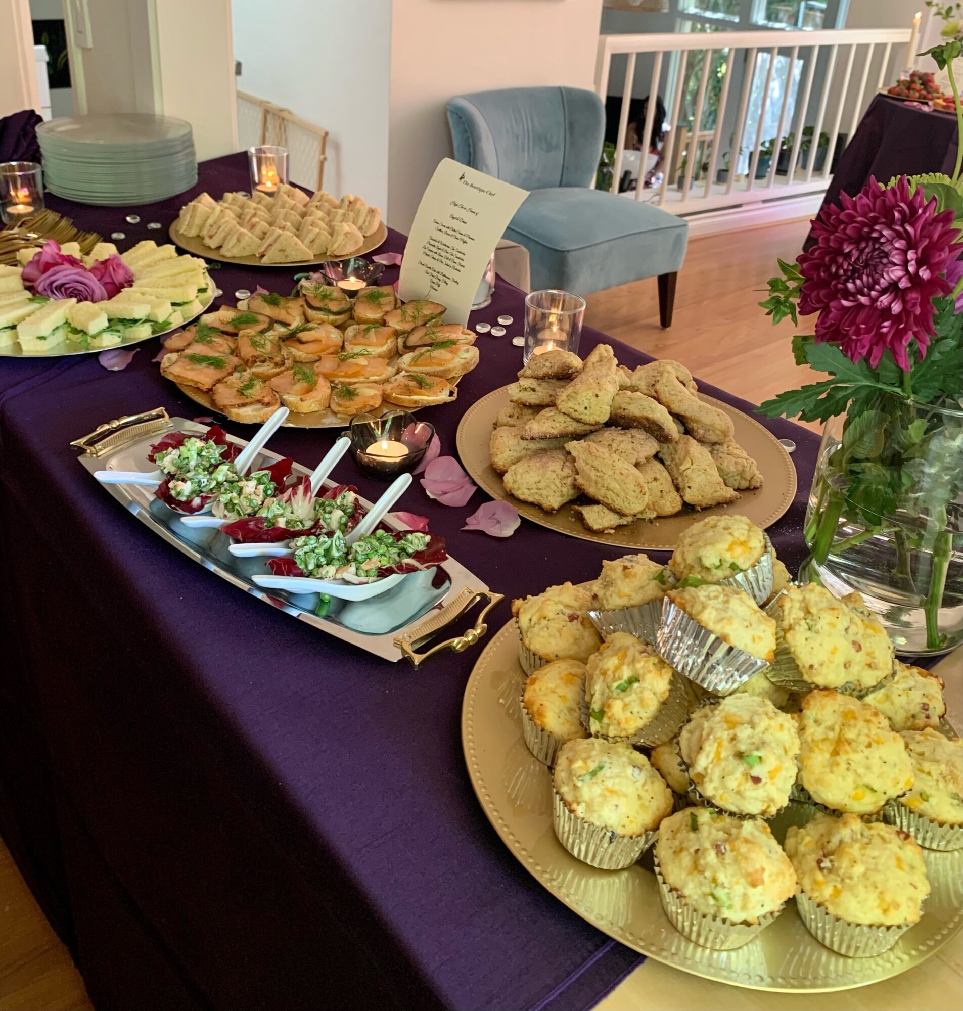 Table with assorted baked goods and snacks, purple tablecloth, flowers, and a seating area in the background.