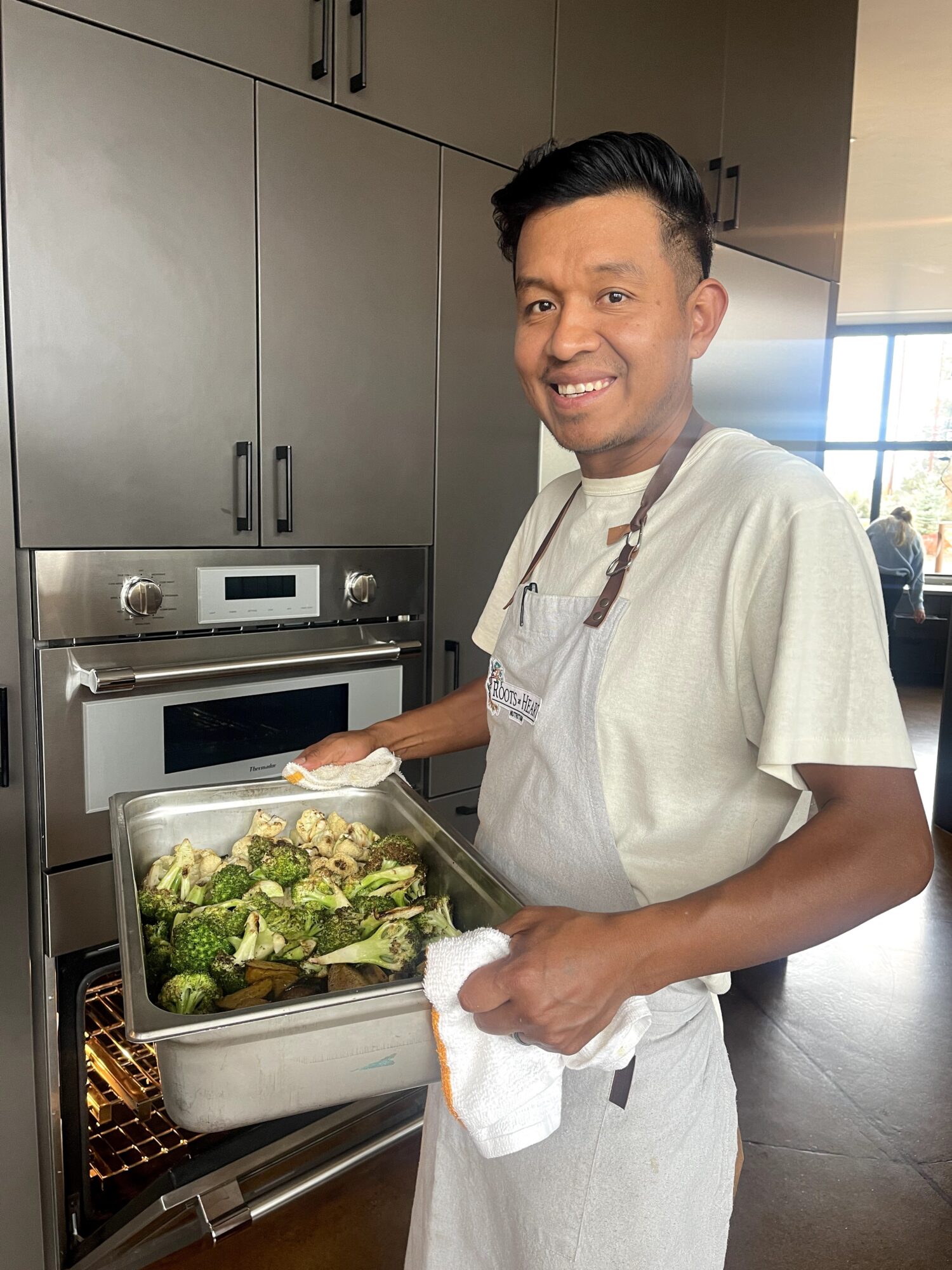 Smiling man holding a tray of food in a modern kitchen, wearing a white apron and casual clothes.