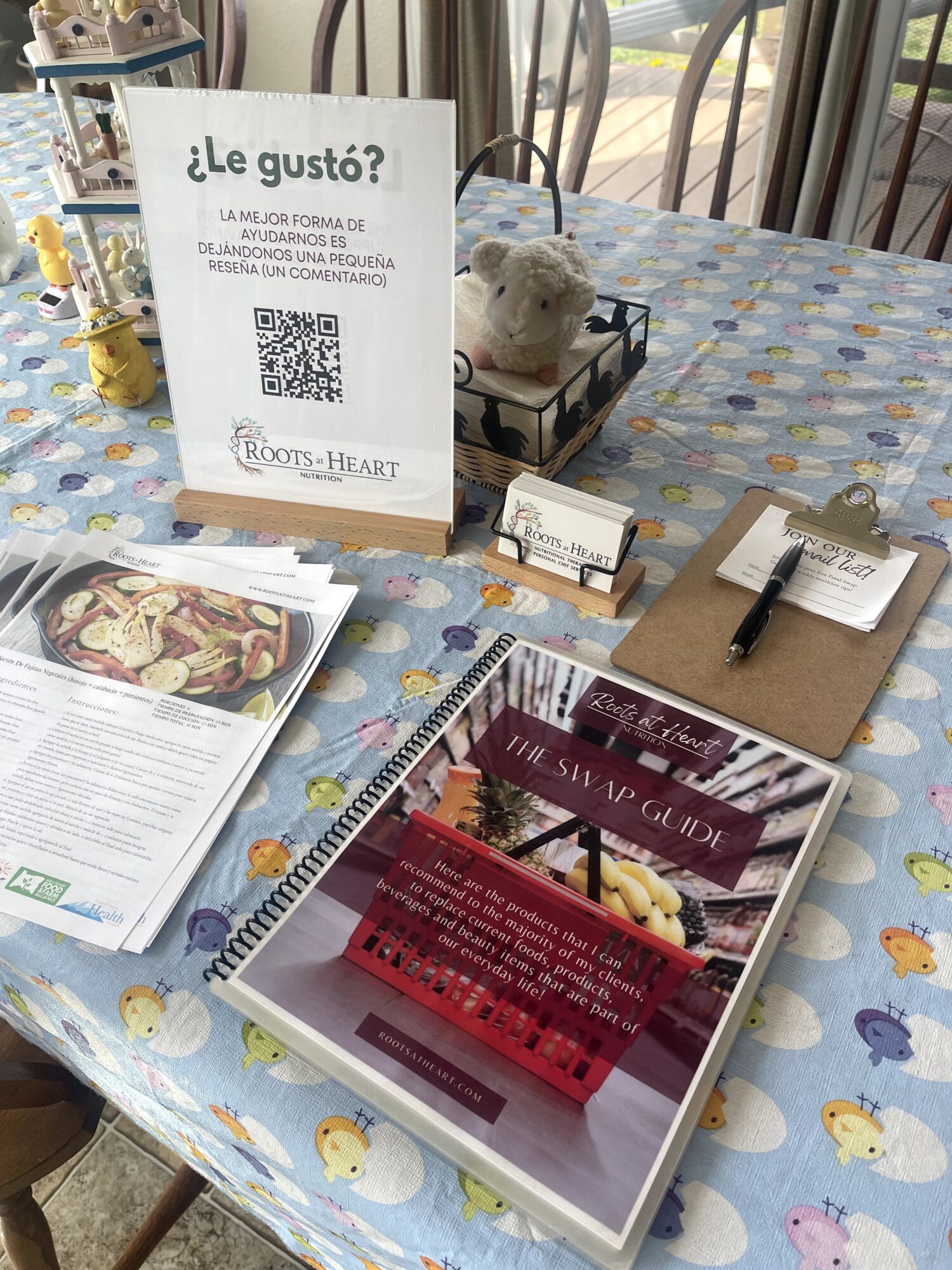 Table with a sign, plush toy, notebook, pen, and a menu on a colorful tablecloth with duck patterns.