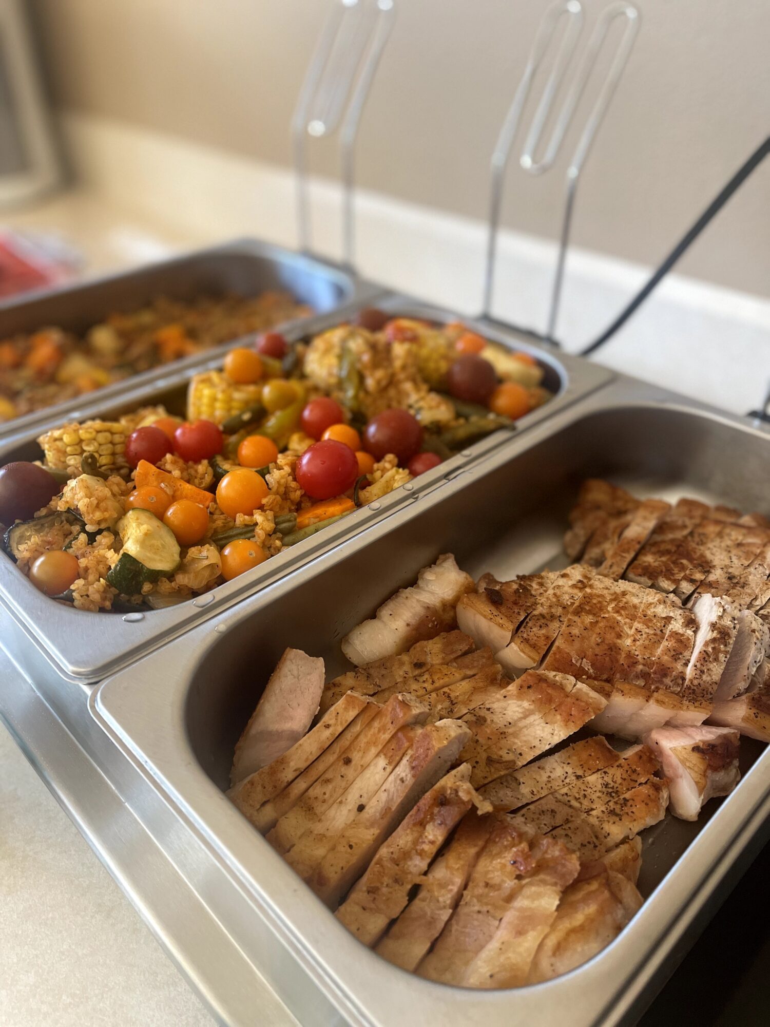 Two metal trays of food on a buffet table, one with sliced cooked meat and the other with mixed vegetables and cherry tomatoes.